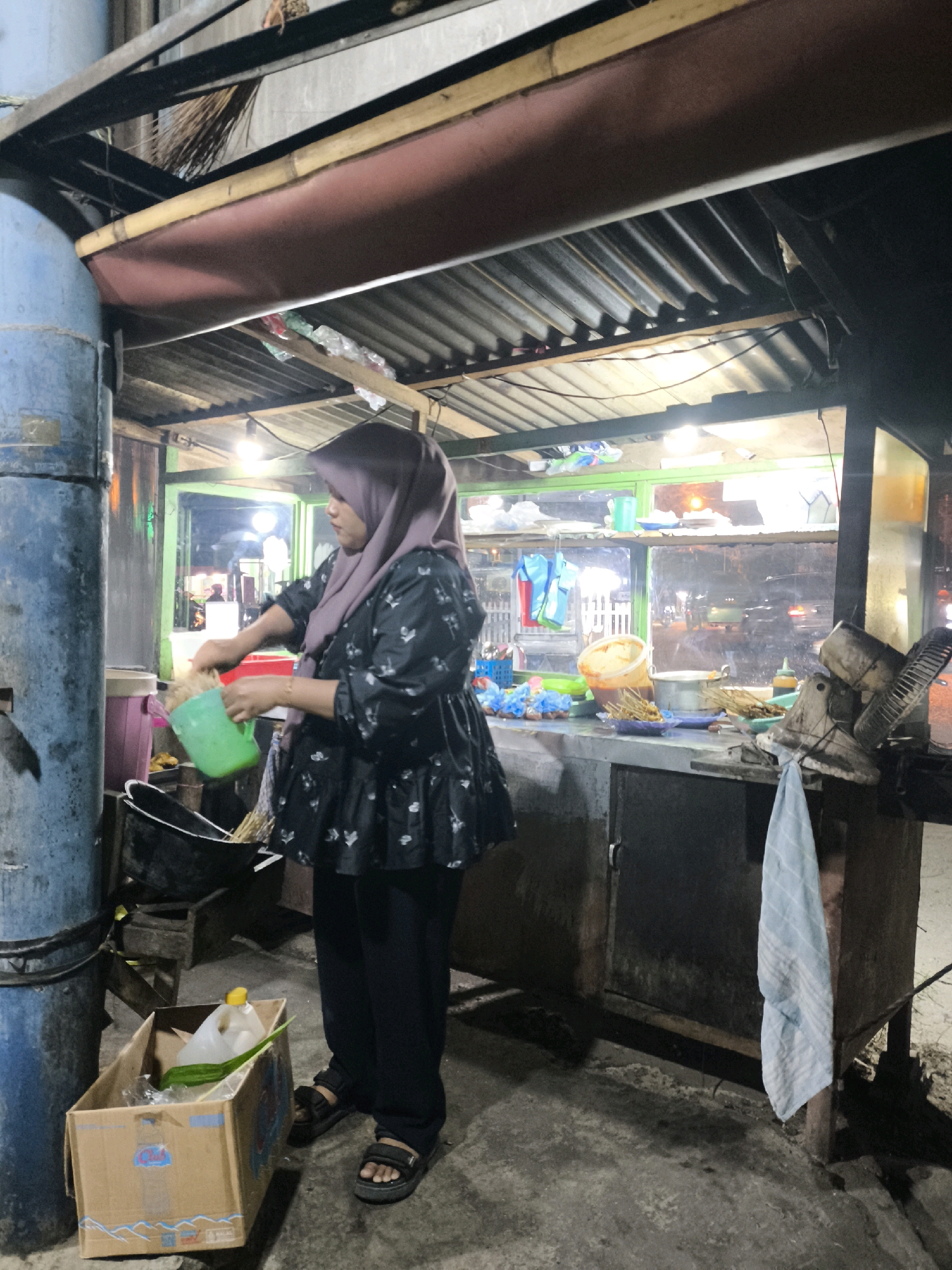 A woman wearing a purple hijab and a patterned black blouse is standing at a food stall in Luwuk, Indonesia. She is pouring something from a green jug into a black pot. The stall appears to be outdoors, with a corrugated metal roof supported by wooden beams. A blue cylindrical object is to the left of the woman, and a cardboard box containing a plastic bottle and a green leaf is on the ground to her right. The background shows a window with various items displayed, including food skewers, plastic bags, and containers. The scene is dimly lit, suggesting it is nighttime or dusk.