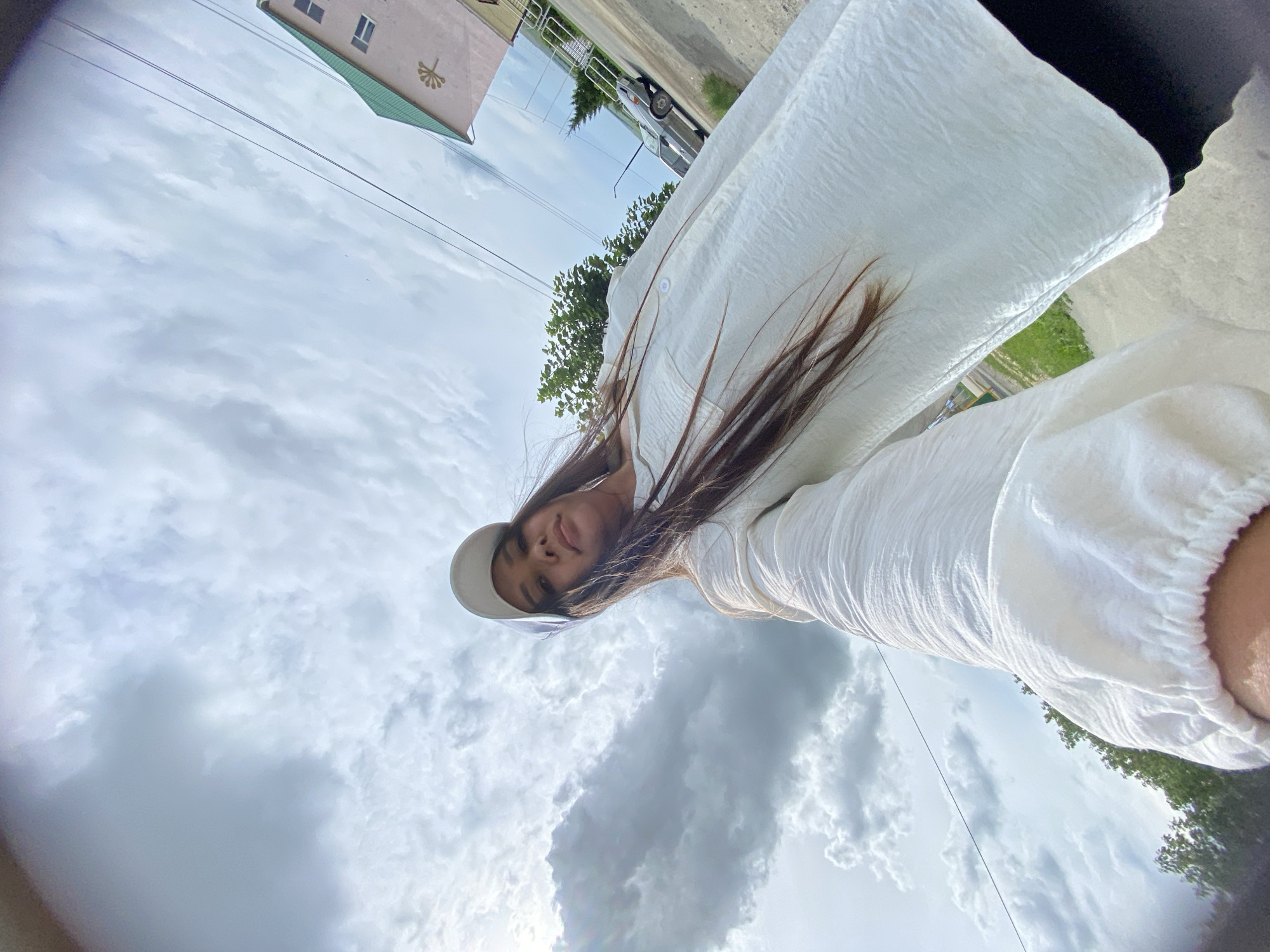 A low-angle, wide-angle shot captures a young woman with long, dark hair wearing a white baseball cap and a white, textured long-sleeved shirt. She is holding the camera out, taking a selfie. The background shows a cloudy sky with dramatic cloud formations, a building with a green roof, power lines, trees, and a road with a car. The location is identified as the city of Akyar, Russia. The woman appears to be outdoors, possibly on a street or pathway.