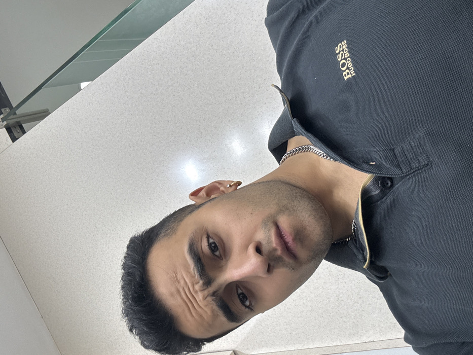 A male individual is depicted in a close-up, angled portrait. He has dark, short hair, furrowed eyebrows, and a light stubble, looking directly at the camera with a serious or questioning expression. He wears a small gold-colored hoop earring in his right ear and a dark-colored polo shirt with a gold "BOSS HUGO BOSS" logo on the left chest. A silver-colored chain necklace is visible around his neck. The shirt's top two buttons are unfastened, revealing a lighter-colored interior placket. The background features a light-colored, textured wall with two visible light reflections. In the upper right, a glass panel secured by metal fixtures is visible, suggesting a modern indoor environment. There are no identifiable visual elements within the image to confirm a specific location within the city of Kolkata, India.