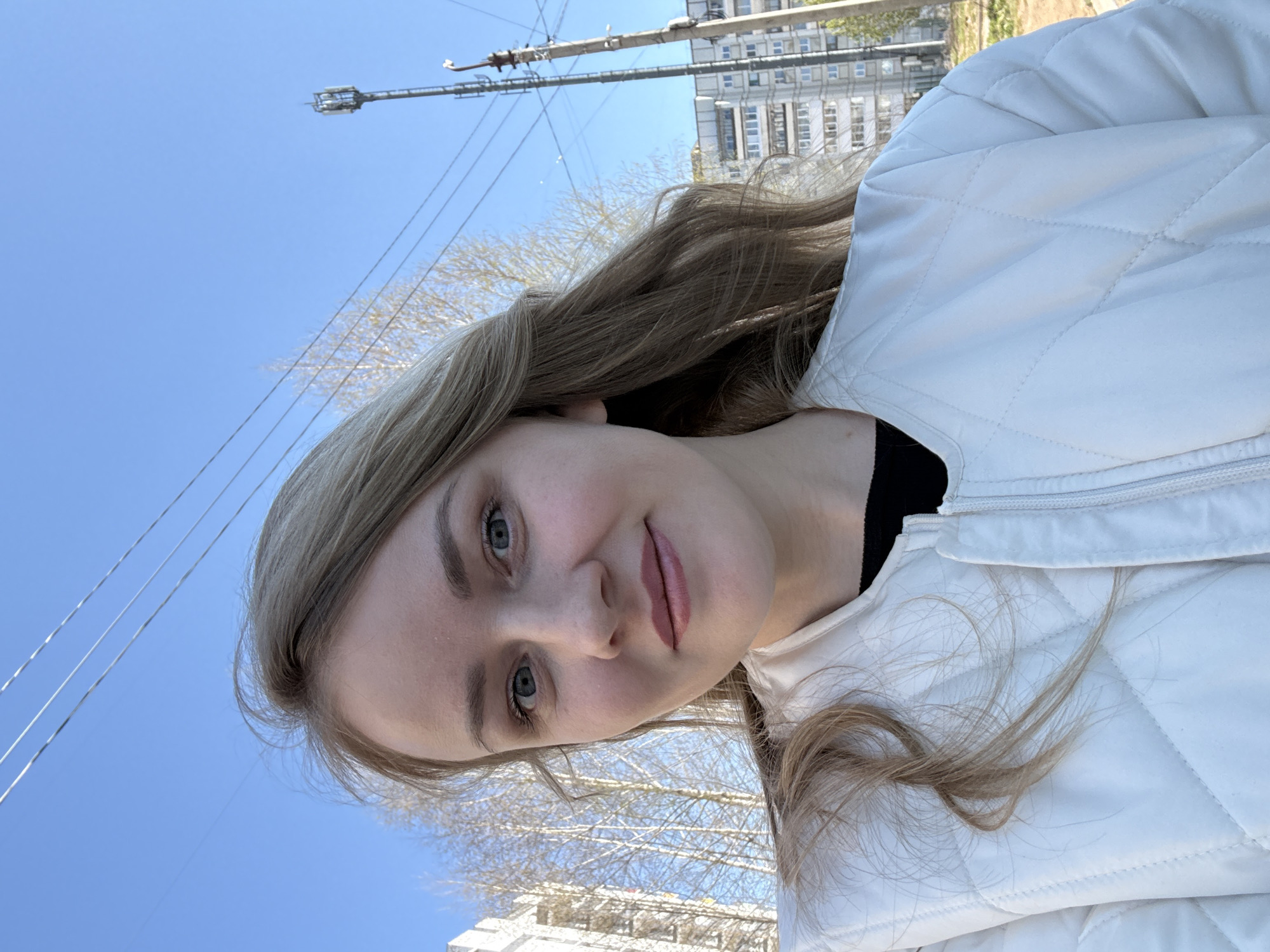 A woman with light brown hair and fair skin is shown in a selfie against a backdrop of a clear blue sky, power lines, a utility pole, bare trees, and a building in the background. She is wearing a white, quilted jacket over a black top. Based on the visible features, the location is speculated to be in the city of Chaikovsky, Russia.