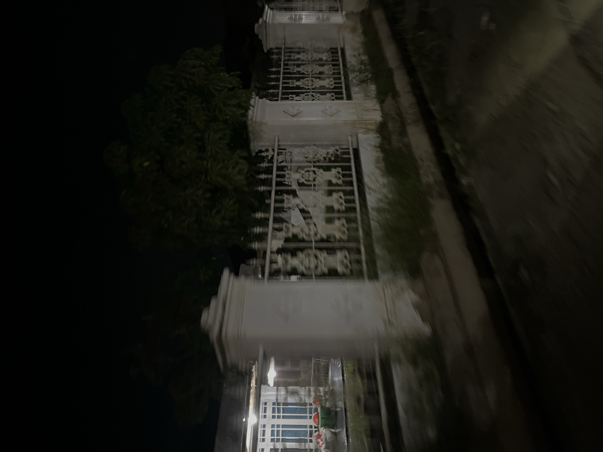 The image depicts a nighttime scene in Kebuntanahterban, Indonesia, showing a blur of motion. In the foreground, a dark, indistinct road or pathway is visible, with a shallow channel running alongside it. Beyond this, a white metal fence with decorative patterns and square pillars marks the boundary of a property. Through the fence, a building with illuminated windows can be seen, suggesting it is a residential or commercial establishment. Some green vegetation, possibly trees or bushes, is present behind the fence and around the pillars. The overall impression is of a moving vehicle capturing a fleeting glimpse of the streetscape at night.