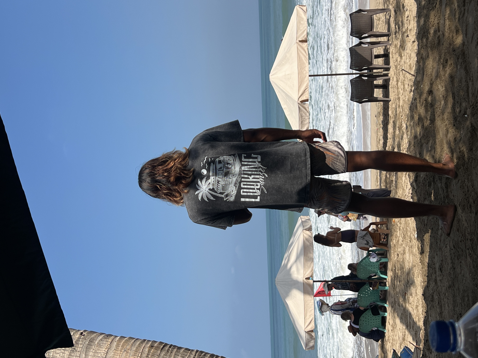 A man stands on a sandy beach in Kuta, Indonesia, facing away from the camera. He is wearing a dark grey t-shirt with a graphic design on the back that appears to depict a camper van and a palm tree, with the text "LOOKING SUNSET" below it. He is also wearing patterned shorts.
Behind the man, the ocean stretches to the horizon under a clear blue sky. Gentle waves are rolling onto the shore. On the beach, two large, light-colored umbrellas are set up, providing shade. Under the left umbrella, several people are gathered, some sitting on green plastic chairs. A red flag is visible near this group. Under the right umbrella, three dark plastic chairs are arranged facing the ocean.
In the foreground, out of focus, is the top of a blue plastic bottle. The overall scene suggests a relaxed day at the beach.