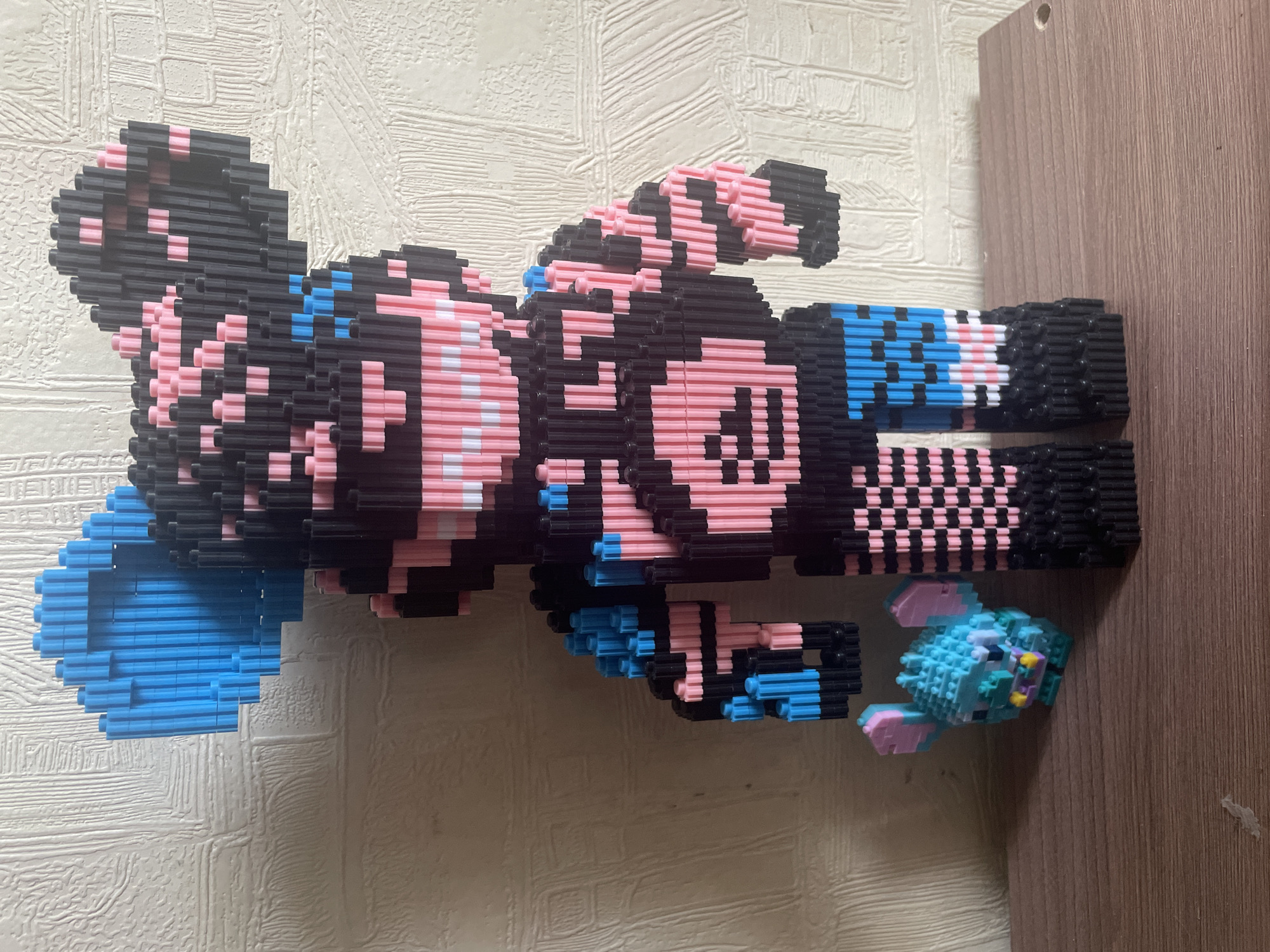 The image captures two pixelated figures, meticulously constructed from small, interlocking building blocks, presented indoors. The dominant subject is a large, stylized character, primarily black and pink, with accents of blue and white. Its design evokes a retro, pixel-art aesthetic, with distinct features forming a head, torso, arms, and legs. One arm extends outwards, while the other is bent.
Positioned near the lower body of the large figure is a smaller, whimsical creature, also made of building blocks. This creature is predominantly light blue and pink, accented with touches of yellow and purple, and appears to be a fantastical animal, possibly aquatic, with fins or wings.
The background consists of two distinct surfaces: a textured, off-white or light beige wall with a linear pattern on the left, and a smooth, light brown wooden surface on the right. A small, circular hole is visible near the top edge of the wooden surface. The figures are static displays, with no activity taking place. The overall image is presented in a sideways orientation. There is no visible text.