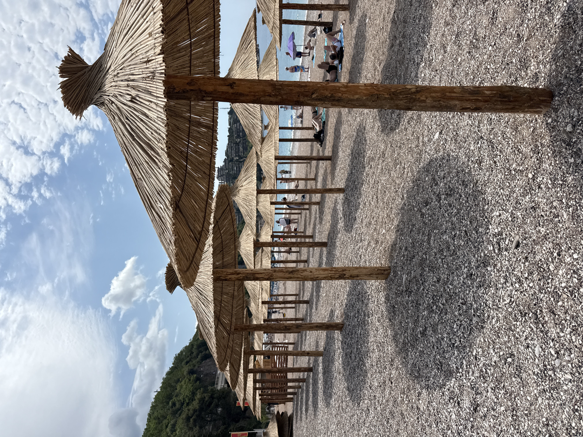 The image captures a picturesque beach scene, viewed along a long, symmetrical row of traditional straw beach umbrellas. The perspective is from beneath the canopy of the closest umbrella, looking out towards the sea. Each umbrella features a thick, thatched top made of natural reeds or dried palm fronds, supported by sturdy wooden poles.
The beach surface consists of small, light-colored pebbles and shell fragments, with distinct shadows cast by the umbrellas indicating bright, direct sunlight, likely during late morning or early afternoon. In the background, numerous people are visible scattered along the shore, engaging in various beach activities such as sunbathing on towels, standing, or walking near the water. One purple beach umbrella stands out amongst the distant figures.
Beyond the beachgoers, the calm, blue sea stretches to the horizon under a clear sky dotted with wispy white clouds. To the left, a lush, green, tree-covered hillside or mountain dominates the landscape, hinting at the rugged coastal geography. The scene evokes a sense of relaxation and vacation. There is no visible text in the image. The setting is characteristic of Mediterranean coastlines, consistent with Budva, Montenegro.