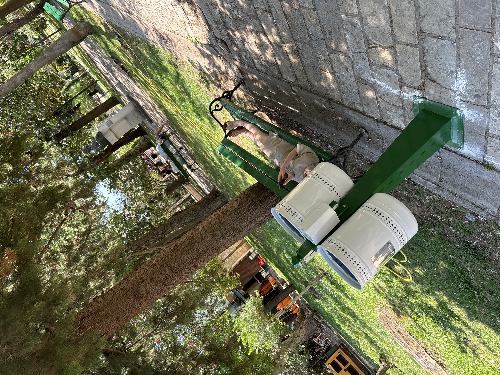 The image depicts an outdoor scene in what appears to be a park or public garden area, likely in Budva, Montenegro, given the provided context. A person is seen relaxing on a green park bench, lying down with their legs extended. They are wearing light-colored clothing and appear to be holding a phone. The bench is situated on a paved pathway, with grass and trees surrounding the area.
In the foreground, two white cylindrical bins with perforated sides are mounted on a green metal structure. One of the bins has a visible label with some text, though it's partially obscured. Behind the bench and bins, a large tree trunk is prominent, with lush green foliage from surrounding trees and bushes. Sunlight filters through the leaves, casting dappled shadows on the ground, suggesting it is daytime, likely late morning or afternoon.
The overall atmosphere of the image is one of leisure and relaxation, with the individual enjoying a quiet moment outdoors. The setting appears well-maintained, with the paved path and manicured grass. In the background, glimpses of buildings and possibly other outdoor furniture can be seen, hinting at a developed area within the park. The presence of the bins suggests a public space designed for visitors.