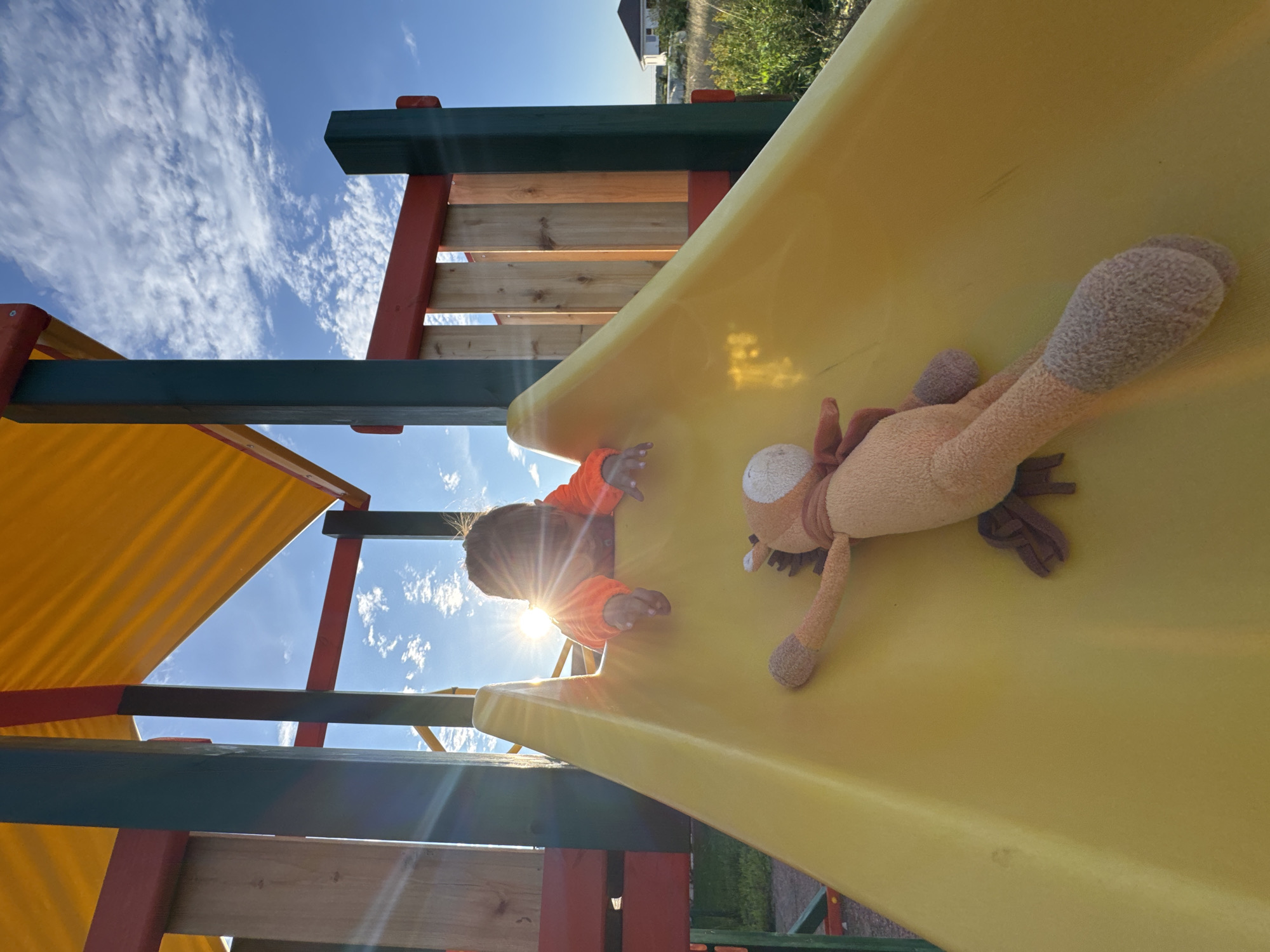 In the city of Kempelevo, Russia, a young child, facing away from the camera, is positioned on the top of a bright yellow slide of a playground structure. The child wears an orange long-sleeved shirt and their head is partially obscured by the sun, which is shining brightly, creating a lens flare effect. A stuffed toy animal, a brown and tan horse with a pink scarf, lies on the slide in front of the child, facing away from them and towards the bottom of the slide. The playground structure is made of wood and metal, with red and green accents. The background features a clear blue sky with scattered white clouds, and a hint of a suburban residential area with houses visible in the distance. The overall scene suggests a sunny day at a playground.