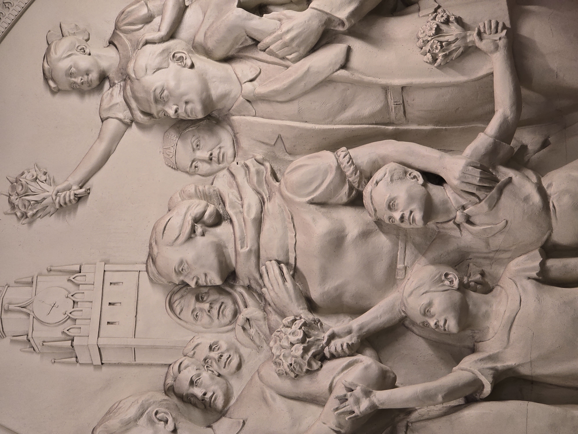 The image shows a bas-relief sculpture depicting a group of figures centered around a larger figure, possibly an adult, surrounded by children. The scene also includes a depiction of a clock tower. The figures are rendered in a pale, possibly plaster-like material, with detailed facial features and clothing. Several of the figures hold bouquets of flowers. The clock tower is depicted with visible clock hands.
This bas-relief is located in Moscow, Russia, and is a memorial to the victims of the 2002 Moscow theater hostage crisis.