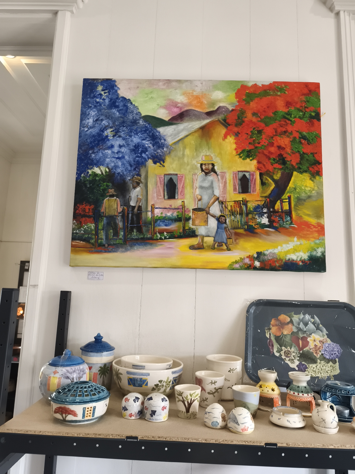 The image shows a still life scene within a gallery or shop. Dominating the upper portion is a colorful oil painting depicting a vibrant outdoor scene with a house, two men by a fence, and a woman and child in the foreground. The painting's style is loose and expressive, with bright blues, reds, yellows, and greens.
Below the painting, a shelf is laden with various ceramic items. These include patterned jars with lids, decorative bowls with what appear to be palm tree motifs, smaller decorative pots, salt and pepper shakers decorated with fish and shells, and candle holders. The ceramics display a variety of colors and artistic styles, some featuring building facades and trees.
To the right of the ceramics, a dark blue decorative tray with a floral pattern is visible. The overall impression is of a curated display of artistic and handcrafted objects. The location, La Plaine-des-Palmistes, France, is noted on a small handwritten tag placed below the painting. The environment appears to be indoors with white paneled walls and simple shelving units.