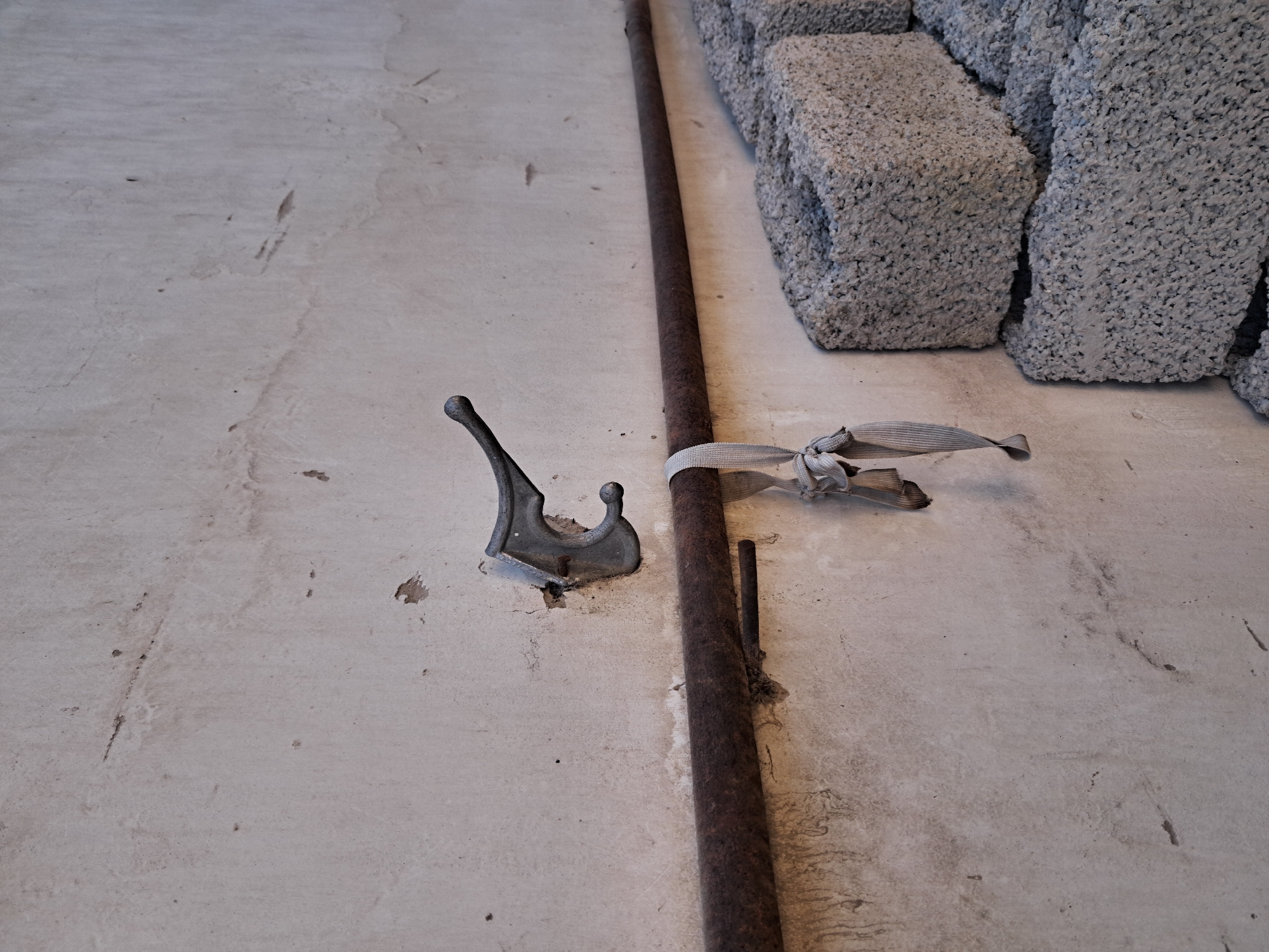 The image depicts a section of a concrete wall in Bagish, Uzbekistan. Mounted on the wall is a weathered metal coat hook. Below the hook, a rusty metal pipe runs horizontally, supporting a knotted, light-colored fabric strap that hangs down. To the right of the pipe, several concrete cinder blocks are stacked against the wall. The wall itself shows signs of age and wear, with minor cracks and stains. The lighting suggests an outdoor environment, likely during daylight. There are no people visible in the frame.