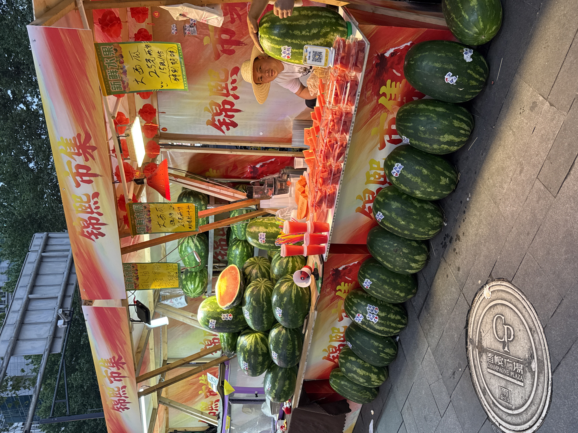 A man in a straw hat is selling watermelons at a fruit stand, located at Champagne Plaza in Jinjiang District, China. The stand has signs advertising "Fresh Fruit Watermelons" for 25 Yuan for two or 15 Yuan for one. He is holding one large watermelon, and there are rows of cut watermelons packaged to sell in the background. There is a megaphone and the stand also has signs with instructions about taking photos. There are lanterns strung on the stand. The ground around the stand is paved.