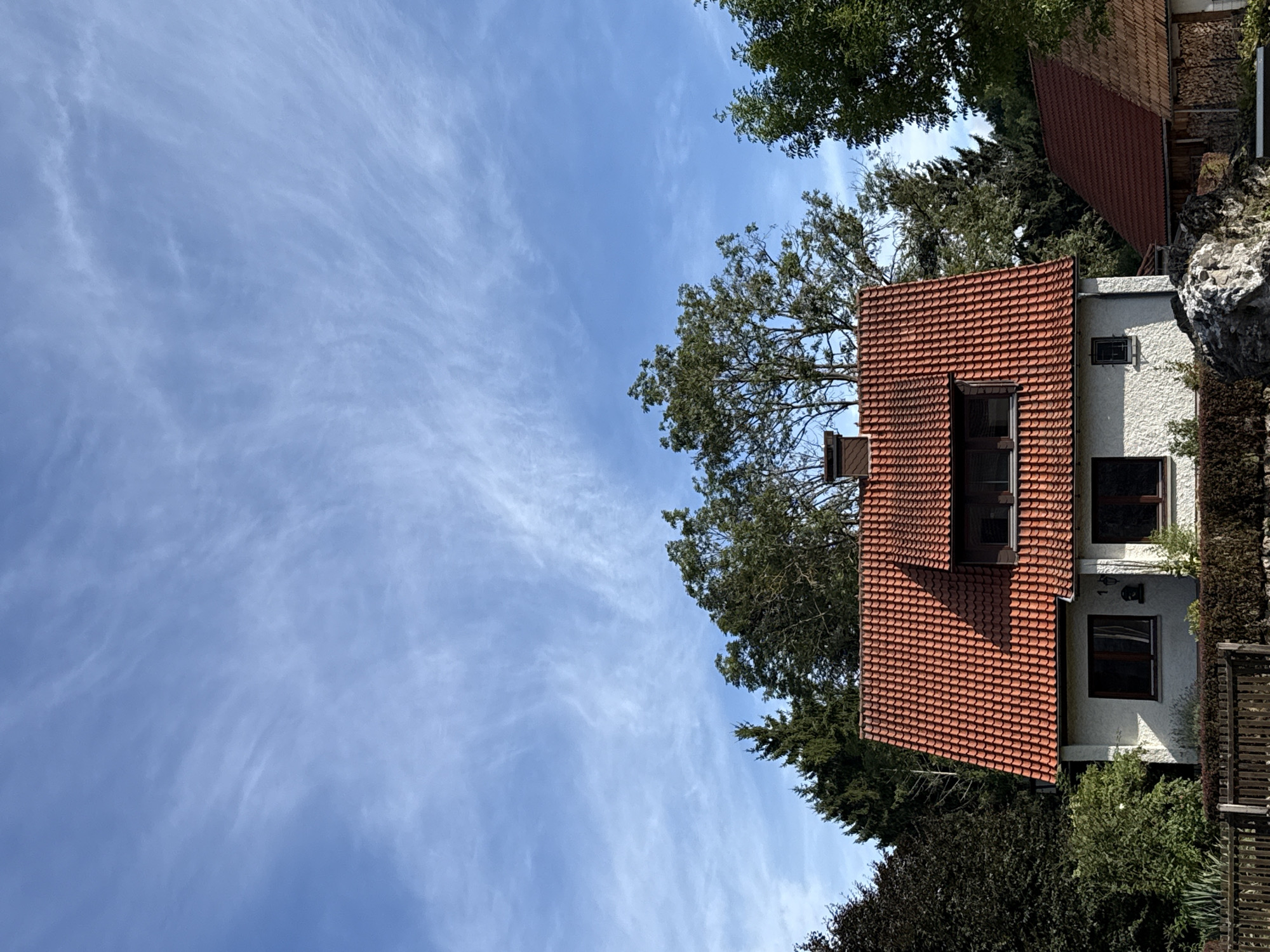The image depicts a residential house with a bright orange, tiled roof in Walkenried, Germany. The house has white stucco walls and dark-framed windows. A dormer window is visible on the front-facing roof section. Trees of varying heights and foliage density surround the house. A wooden fence runs along the front of the property. The sky above is a clear blue with wispy white clouds. The scene appears to be a daytime, outdoor shot, with no people or animals visible.