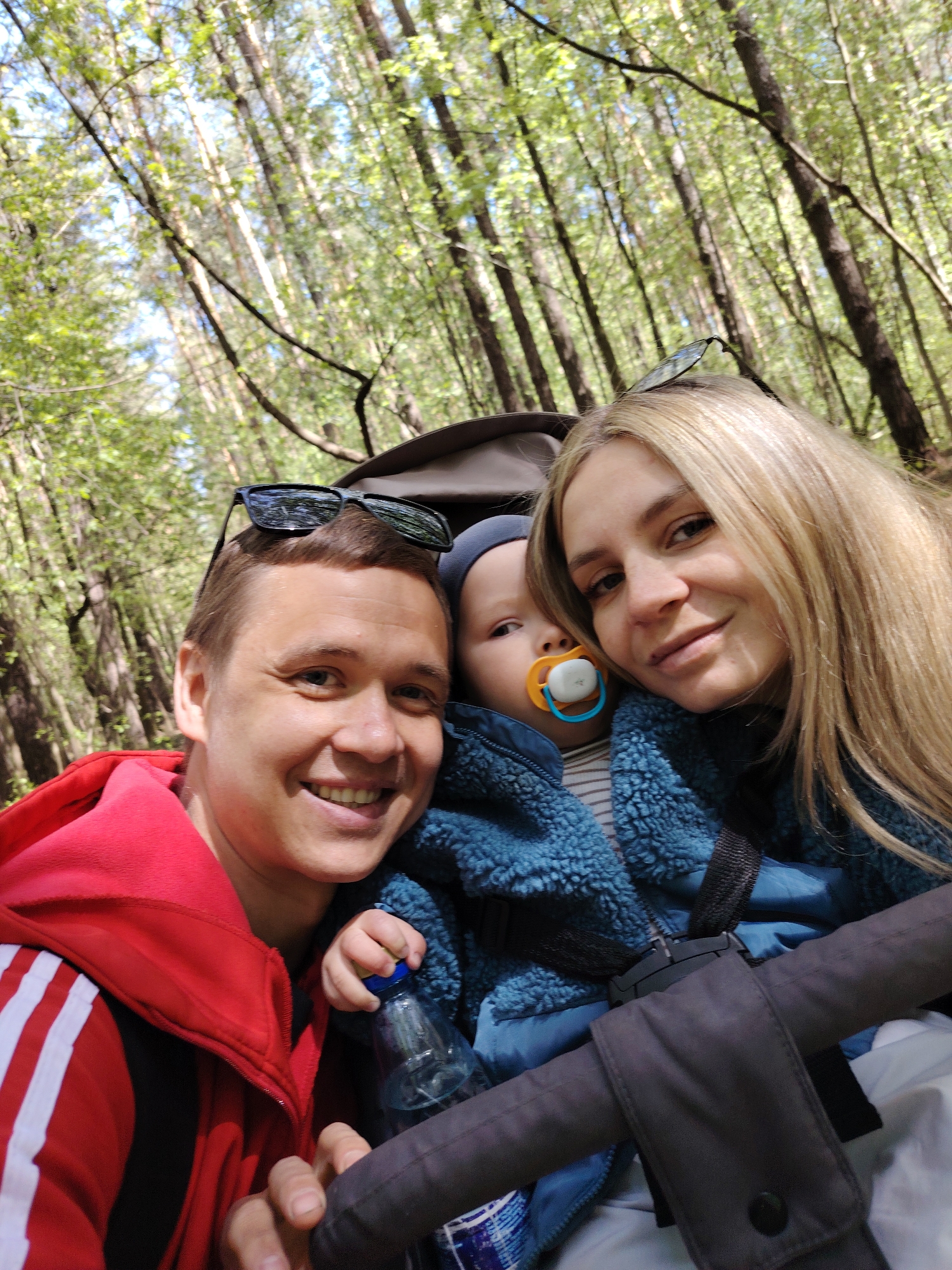 The image shows a family of three in a forest setting. The location is likely within or near the city of Chaikovsky, Russia, given the prompt’s context. A man wearing a red hooded jacket is on the left side of the frame, smiling at the camera. A woman with long blonde hair is on the right, also smiling. Between them sits a baby with a pacifier, bundled in a blue fuzzy jacket. The baby is in a stroller, the frame of which is visible in the foreground. A water bottle is visible in the baby's hand. Sunglasses are perched on top of the man and woman’s heads. The background consists of tall trees with green foliage, indicating a forest environment. The lighting suggests a sunny day.