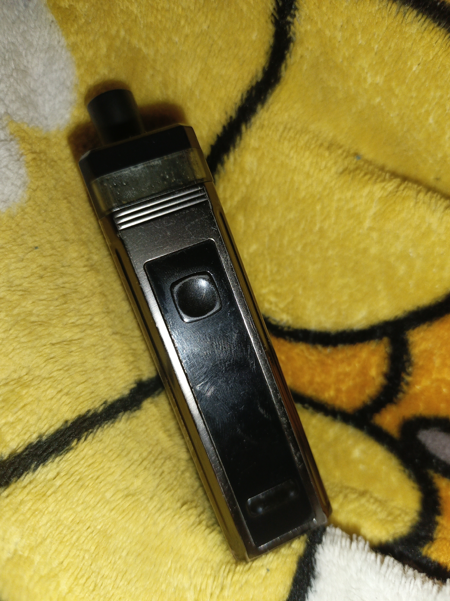 A black, metallic vape pen lies on a yellow and orange patterned fabric. The vape pen is oriented vertically, with the mouthpiece at the top. A black button is visible on the front of the device. The fabric appears to be a soft material, possibly a blanket or towel, with a fuzzy texture. The background is out of focus, suggesting a shallow depth of field. There are no people or other objects clearly visible in the image. There is no information in the image to suggest a location in Shumerlya, Russia.