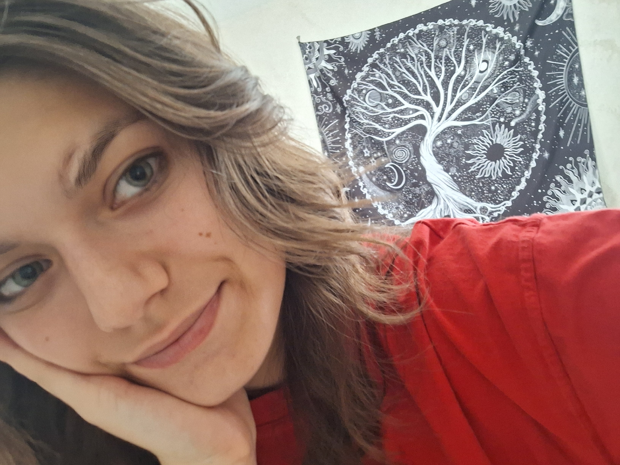 A selfie of a young woman with light brown hair and fair skin, resting her cheek on her hand. She is smiling slightly. She is wearing a red shirt. Behind her is a wall hanging with a tree of life design surrounded by celestial bodies in black and white. The location is likely indoors, but the image doesn't provide enough information to determine a specific location in Panagyurishte, Bulgaria, or any other city.