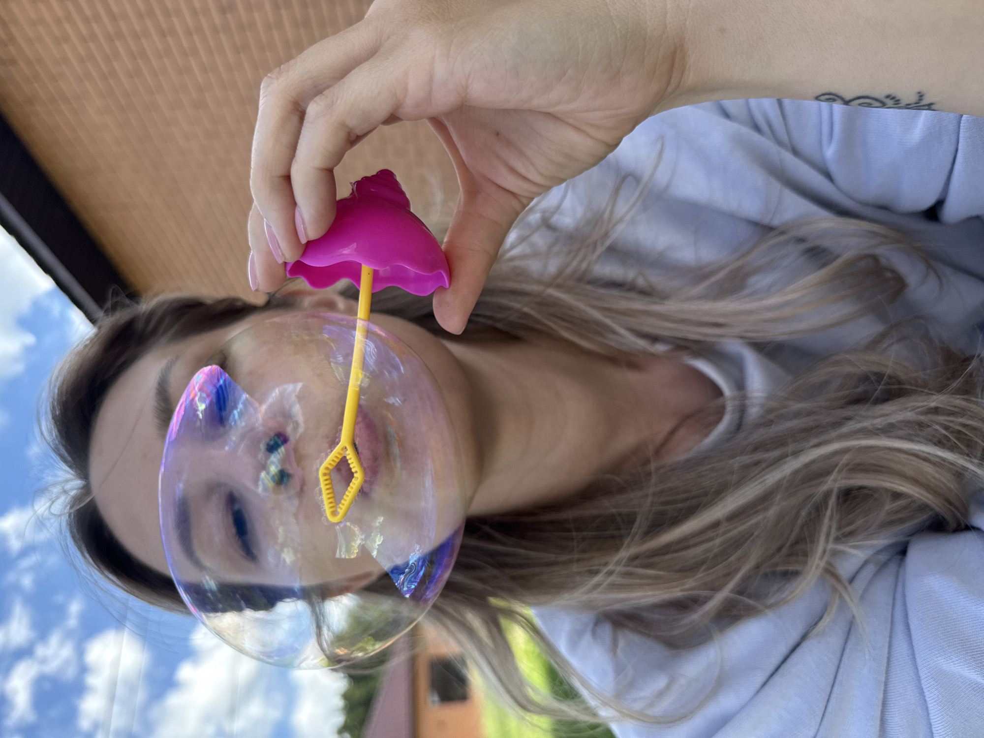 A medium close-up shot shows a young woman blowing a large iridescent bubble outside. The woman, facing the camera with her eyes closed, has blonde hair and is wearing a light blue hoodie. She holds a yellow bubble wand with a pink plastic flower-shaped handle to her lips, from which a large bubble is emerging, distorting her face. The background is an outdoor scene with a brick building on the right, green grass, trees, and a blue sky with white clouds. The location is identified as Chaikovsky, Russia.