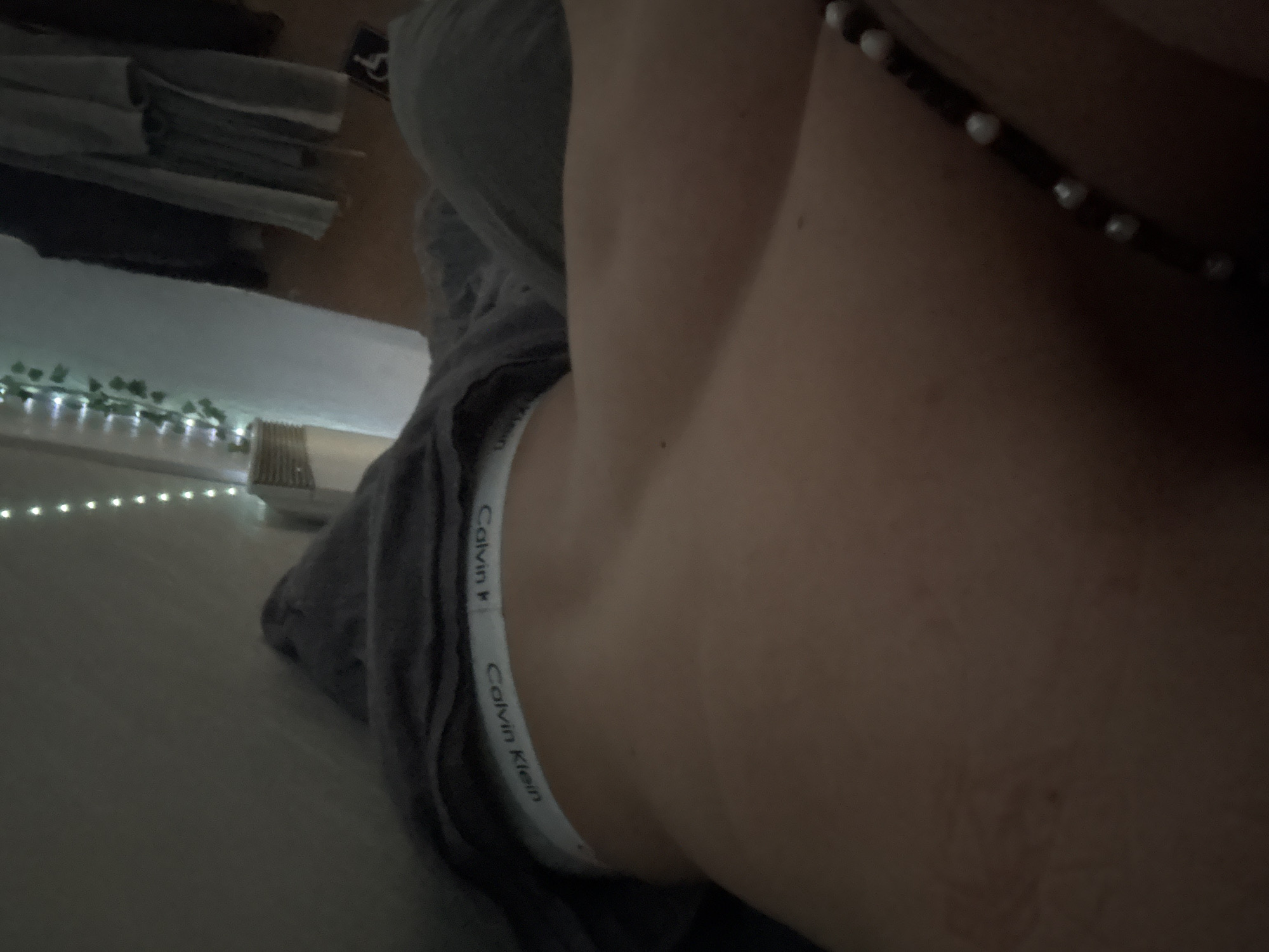 The image presents a close-up view of a person's body, primarily focusing on the waist and hip area. The subject is wearing grey sweatpants and Calvin Klein underwear. A necklace with white beads is visible across the upper portion of the frame. To the left, there is a glimpse of a bed with string lights and stacked folded clothes. The location is not discernible from the provided visual information. Therefore, there is no evidence to suggest this image was taken in Troyan, Bulgaria.