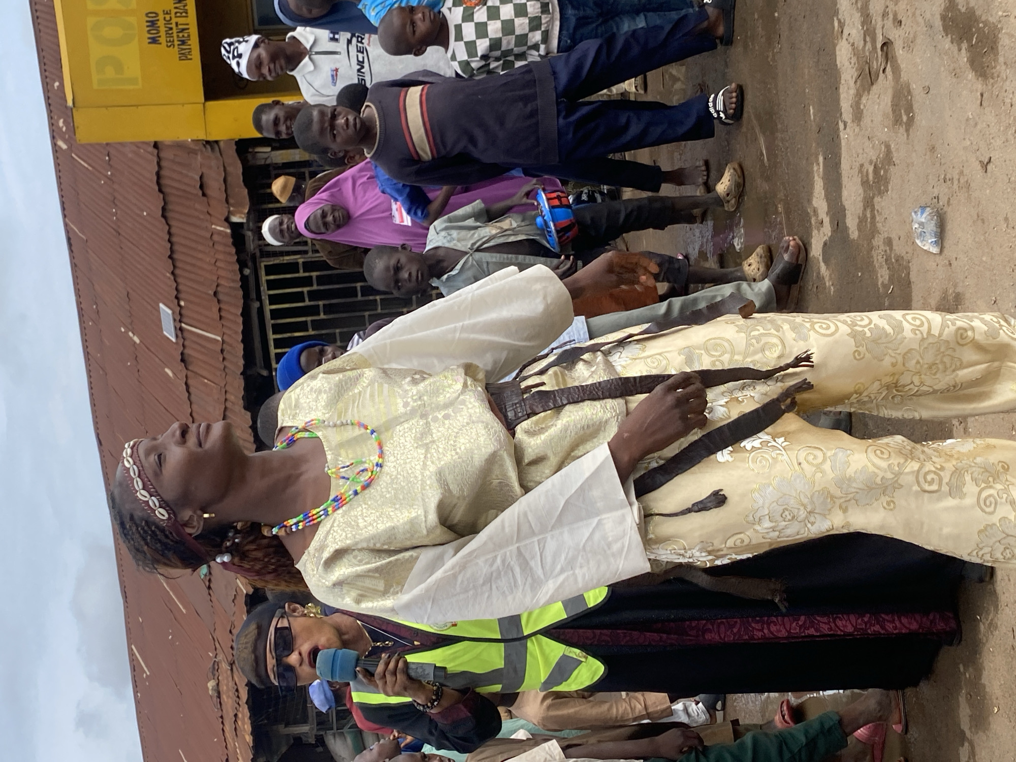 In Rigachikun, Nigeria, a woman in a patterned gold outfit and beaded necklace stands center frame, holding a microphone and wearing a bright yellow safety vest over a black and maroon garment. She is looking to her right with a determined expression, her hair braided and adorned with a seashell headband. Around her, a crowd of people, including many children, gather. Some are looking at the woman, while others are looking elsewhere. The background shows a tin-roofed building with a yellow facade, likely a commercial establishment, with faded lettering that appears to say "POST MOMO SERVICE PAYMENT BANK." The ground is uneven and appears to be wet in places, suggesting recent rain. The overall scene depicts a lively outdoor gathering, possibly an event or a public address, in a bustling Nigerian community.