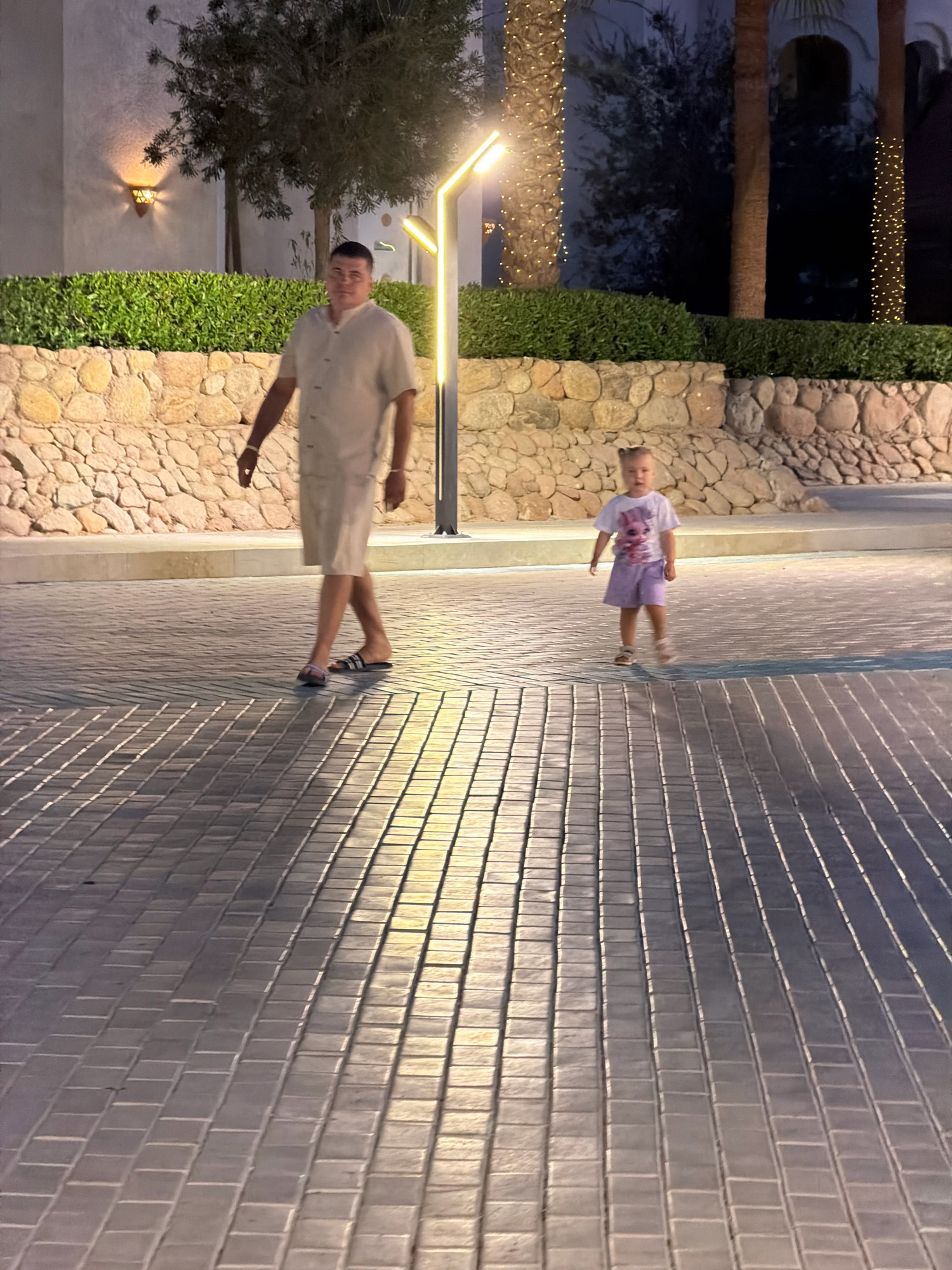An adult male wearing a light-colored button-down shirt and shorts walks on a paved walkway. Beside him, a young girl in a white t-shirt and light purple shorts walks in the same direction. The setting appears to be an outdoor area, possibly a resort, in Sharm El Sheikh, Egypt, with a stone wall and lush green bushes in the background, illuminated by ambient lighting and decorative string lights on palm trees.