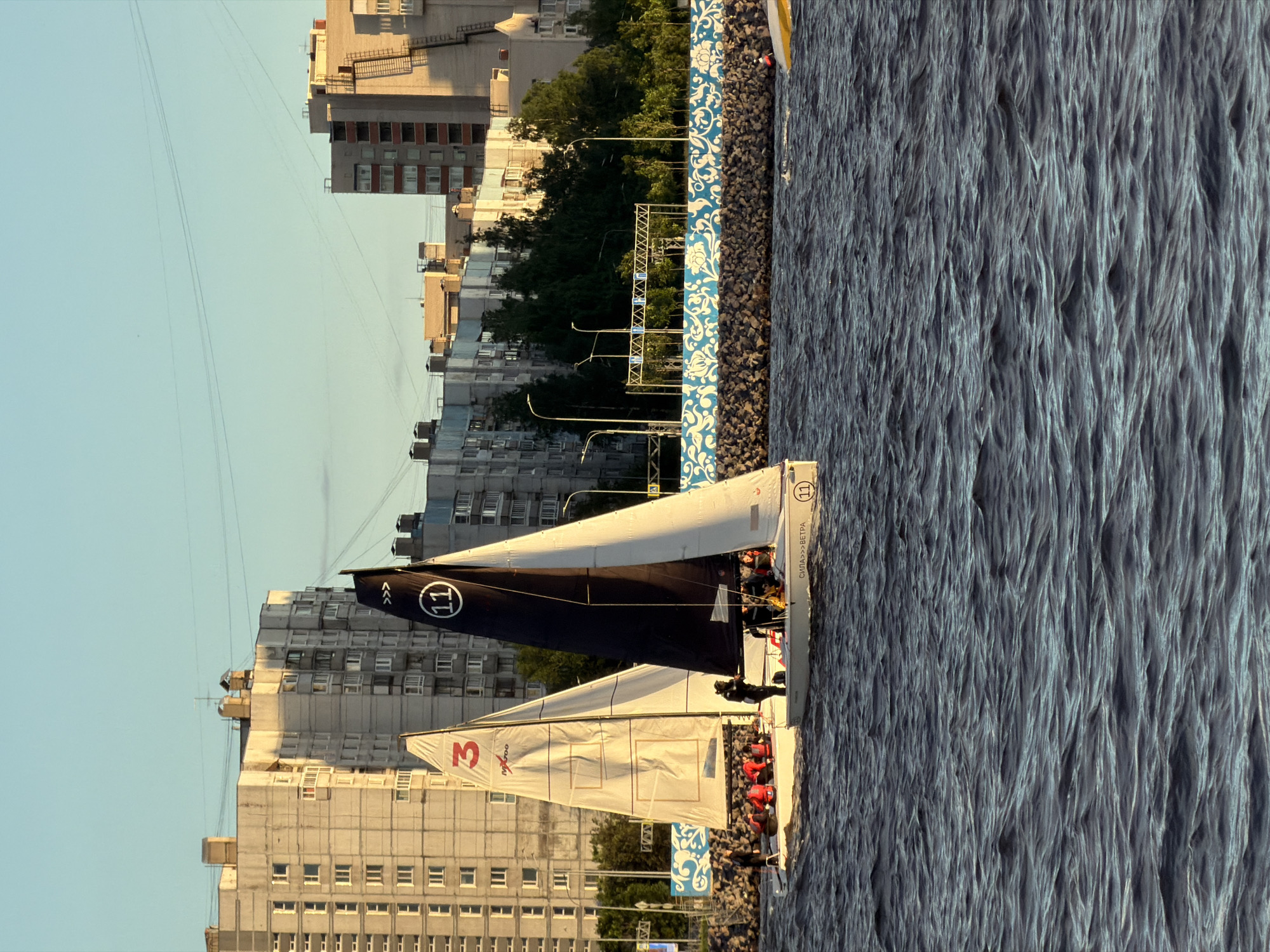The image depicts a fleet of sailboats on the Neva River in Saint Petersburg, Russia, with an urban backdrop of apartment buildings. The sun is low in the sky, casting a warm golden light, suggesting it is either early morning or late evening. Two sailboats are prominently featured in the foreground, their sails filled with wind. One sail is white with a red number "3" and a logo, while the other is black with a white number "11" and lettering that appears to say "СИЛА>>>ВЕТРА", a Russian phrase meaning "Power of the Wind". Both boats have several people on board, some wearing red life vests.
In the background, a row of multi-story apartment buildings line the riverbank. The architecture suggests the Soviet era, with repetitive window patterns and block-like structures. Behind the buildings, a clear sky with a few thin power lines stretches across the horizon. A decorative blue and white patterned wall or fence runs along the riverbank, separating the water from the embankment. The water itself is choppy, with visible waves, indicating a breezy condition. The scene captures a moment of outdoor activity and urban landscape, bathed in the soft glow of sunset or sunrise.
