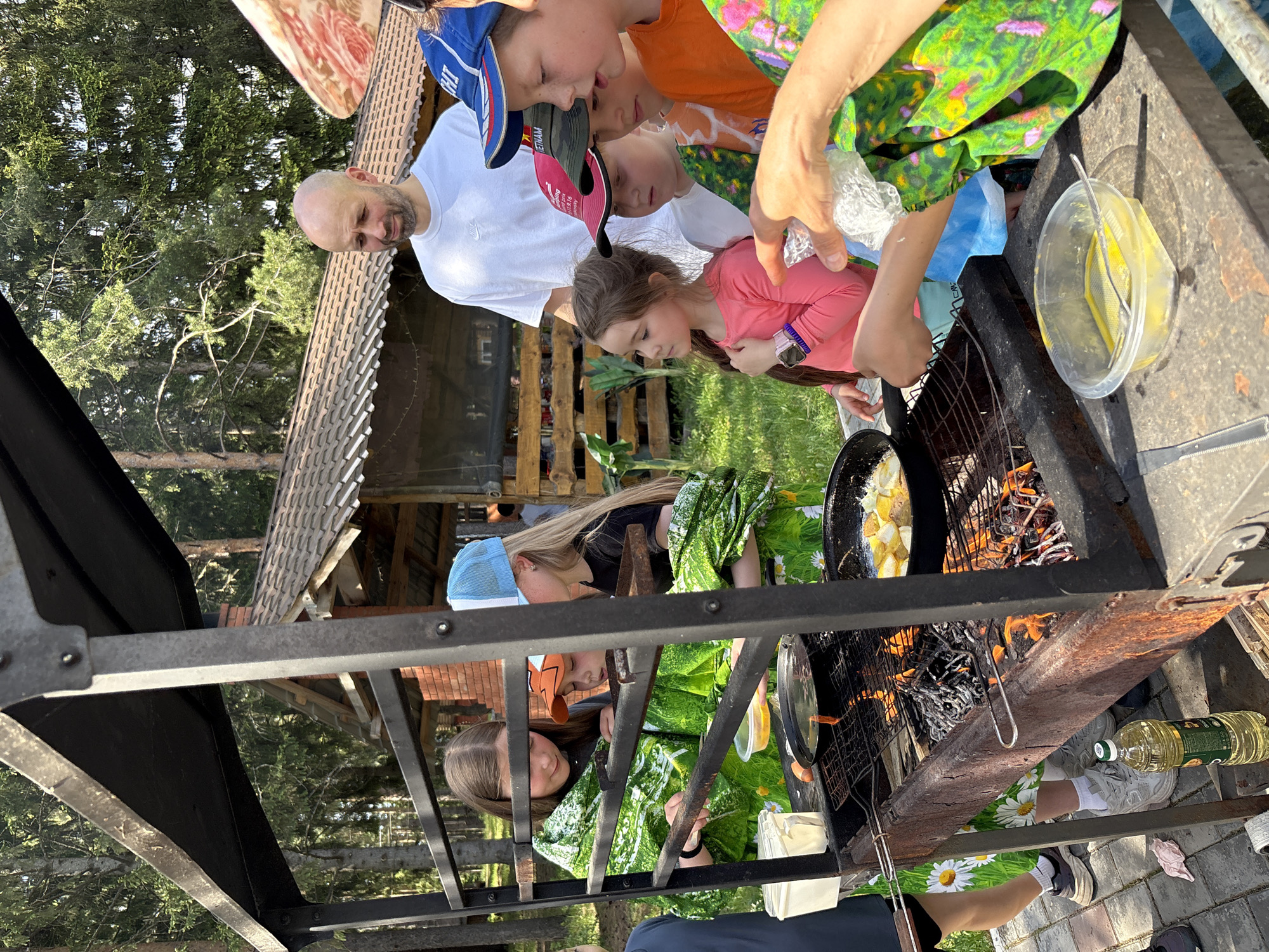 People are gathered around a barbeque pit in an outdoor setting. There are children and adults present, and they are cooking food in a frying pan over an open fire. A container with yellow food is visible next to the grill. A bottle of oil is on the ground near the barbeque pit. The surrounding environment includes trees and a wooden structure in the background. The image was taken in Прикамский, Russia.
