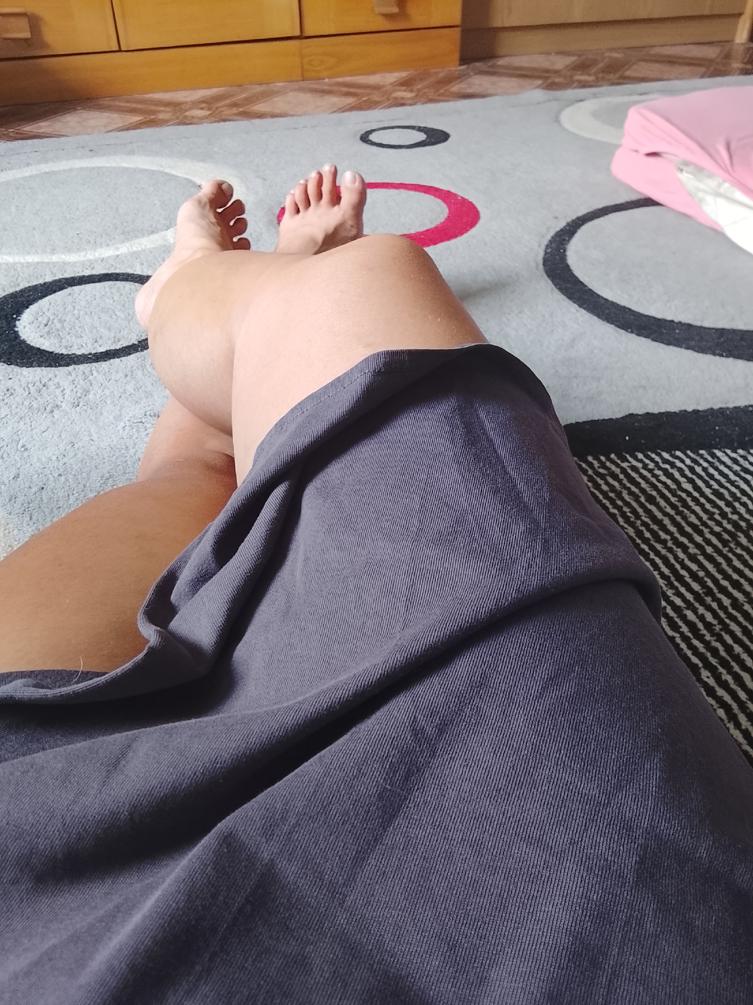 This is a close-up, first-person perspective shot of a person's legs and feet, resting on a patterned carpet. The person is wearing a dark grey skirt or dress, which is draped over their lap. Their bare feet are visible, with toes spread slightly.
The scene appears to be indoors, likely a living space. The carpet has a design of concentric circles in black, white, and red. In the background, a wooden cabinet or dresser can be seen, suggesting a domestic setting. To the right, a pink cushioned object, possibly a pillow or cushion, is partially visible.
The lighting suggests it might be daytime, with natural light filtering into the room. There are no explicit cues about the weather, but the indoor setting implies a comfortable environment. The overall mood is relaxed and casual, as indicated by the pose and the domestic setting. There is no visible text in the image. The location context of Almaty, Kazakhstan, does not provide any specific visual cues within the image itself.