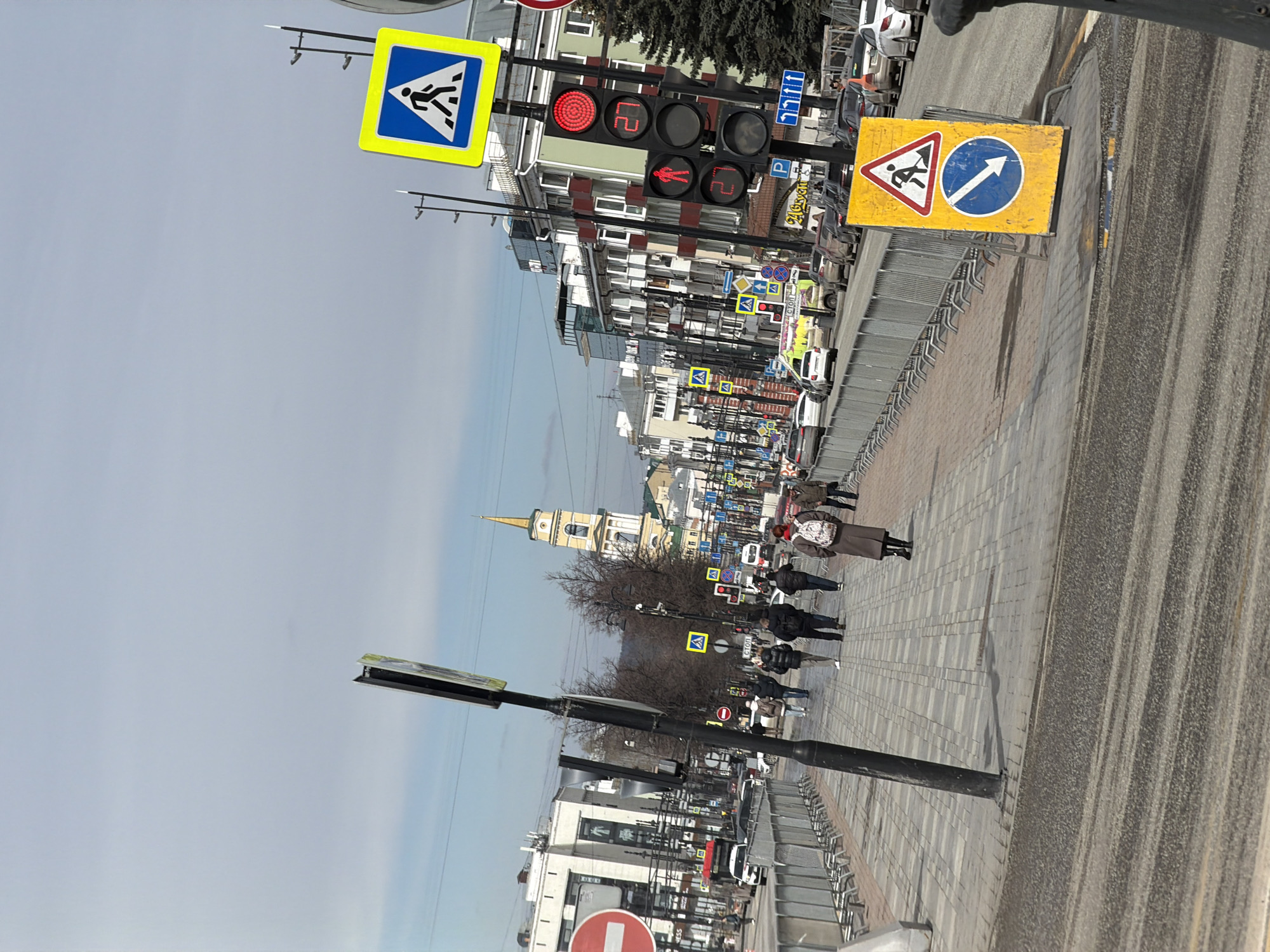 The image depicts a street scene in Perm, Russia, characterized by pedestrian activity and urban infrastructure. Several individuals are walking on the sidewalk adjacent to a roadway with visible vehicles. Traffic signals are present, displaying red lights for both vehicle and pedestrian crossings, which may indicate that pedestrians are intended to cross the street. Numerous street and traffic signs are observable along the street. In the background, a church steeple rises into the sky, providing a distinguishing landmark of the location. The photograph is taken during the day under a sky that appears overcast or partially cloudy. The urban landscape is densely populated with buildings of various heights, suggesting a central or well-developed area within Perm.