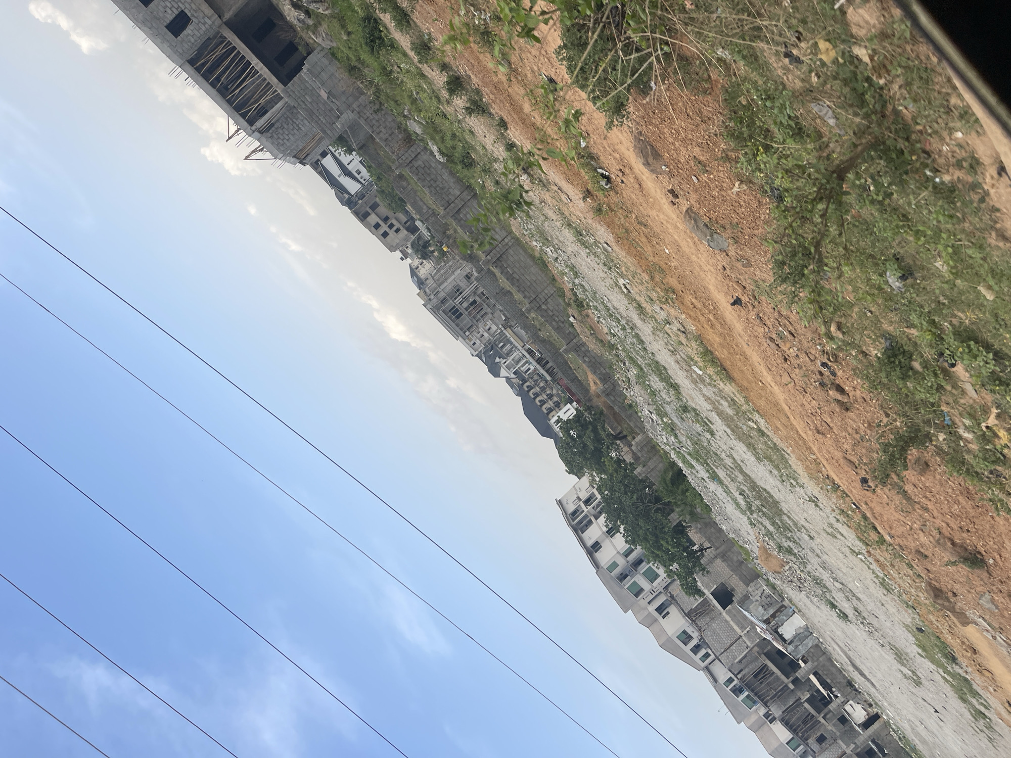 The image depicts a sprawling urban development scene in Abuja, Nigeria, under a bright, partly cloudy daytime sky. The landscape features a mix of ongoing construction, undeveloped land, and established buildings, conveying a sense of growth and transformation.
In the midground and background, numerous multi-story buildings are visible, many in various stages of construction. These structures are built with concrete blocks, some displaying exposed wooden scaffolding and rebar, indicating active development. The architectural style suggests residential or commercial properties. Several completed or near-complete buildings with multiple windows and modern facades are also present.
The foreground and unbuilt areas consist of uneven, reddish-brown earth, characteristic of natural soil. Patches of green vegetation, including small trees and shrubs, are scattered across the ground, interspersed with areas of gravel and construction debris. Some litter is visible among the vegetation and on the ground, suggesting a developing environment.
The sky, occupying the upper portion of the frame, is a clear light blue with scattered, soft white clouds. Multiple dark, thin lines, identifiable as overhead power lines, traverse the sky, implying existing utility infrastructure. No people are visible in the scene, and there is no discernible text. The overall impression is one of active urban expansion and land development.