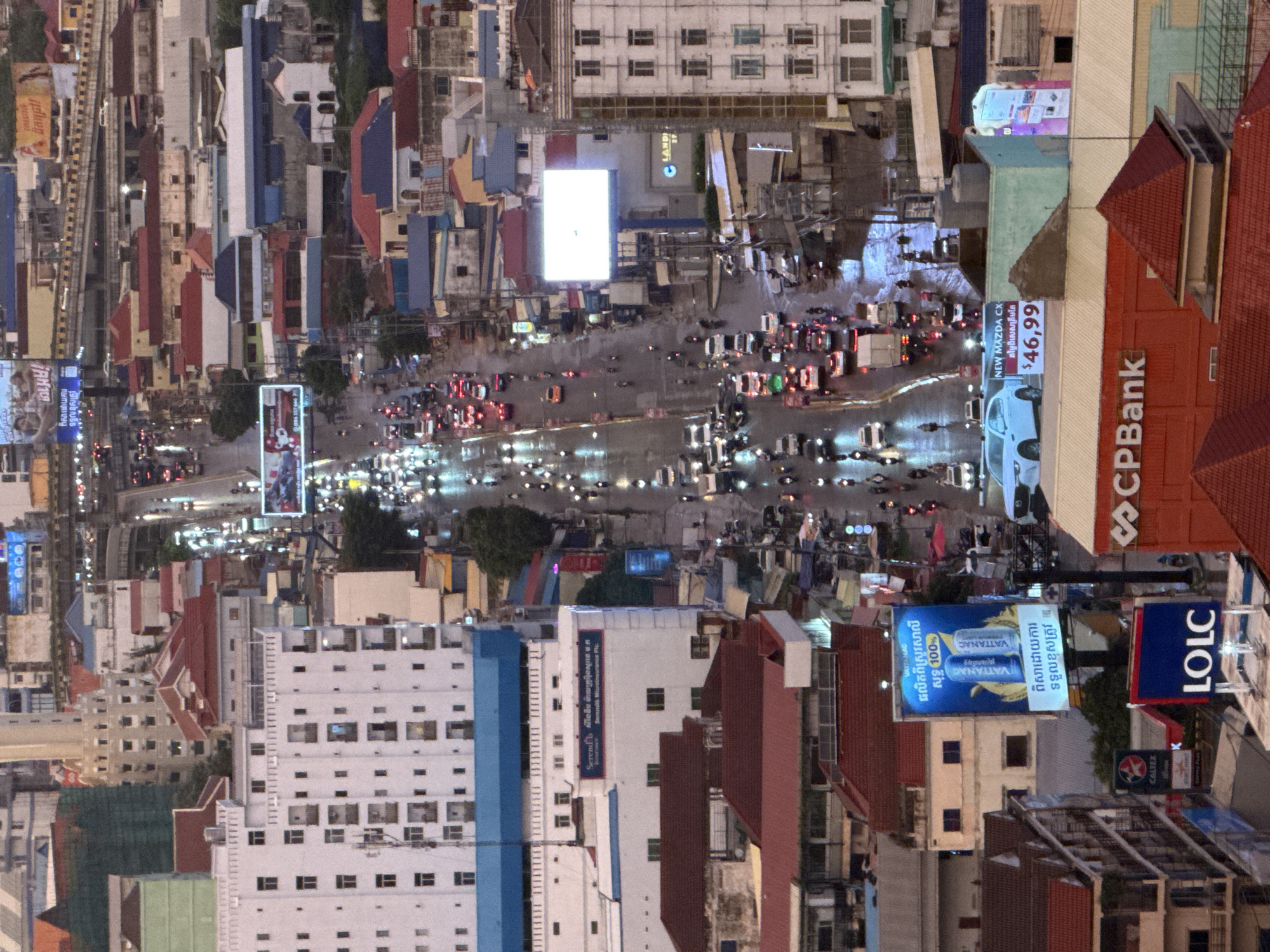 This is a high-angle, wide shot of a busy street in Khan Mean Chey, Cambodia, likely at dusk or during a rainy period, given the wet road surfaces and reflections. The scene is dominated by a multi-lane road choked with traffic, including cars, trucks, and numerous motorcycles. Buildings of varying heights and architectural styles line both sides of the street, some appearing modern and commercial, while others are older residential structures with red tile roofs.
Several large billboards are visible, advertising products and services. One prominent billboard features two cans of "VATTANAC" energy drink with the text "100%" and other Cambodian script. Another billboard displays a white car with the text "NEW MAZDA CX" and a price of "$46,99". A building with the logo "CPBank" is prominent in the foreground. Other signs include "LCLC" and "CALTEX". The streetlights and vehicle lights create a glittering effect on the wet asphalt. The environment appears to be a densely populated urban area. There are no clearly discernible individuals in focus, but the presence of many vehicles implies a significant human population and activity. The sky is overcast, contributing to the muted lighting.