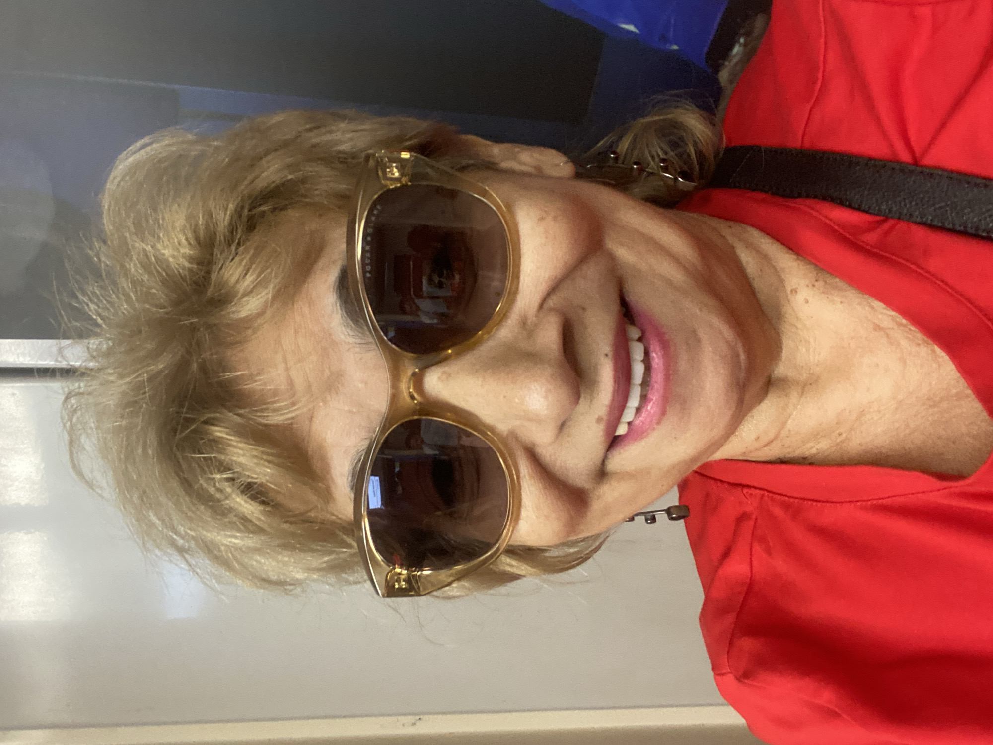 The image shows a close-up selfie of a woman with blonde, feathered hair, wearing large, amber-rimmed sunglasses and a vibrant red top. She is smiling, revealing her teeth, and has a dark strap visible across her shoulder, suggesting a bag is being worn. She is also wearing a dangling earring on her left side. The background is out of focus, appearing to be an indoor setting with a light-colored wall on the left and a darker surface on the right. There is no identifiable Moscow, Russia-specific context in the image.