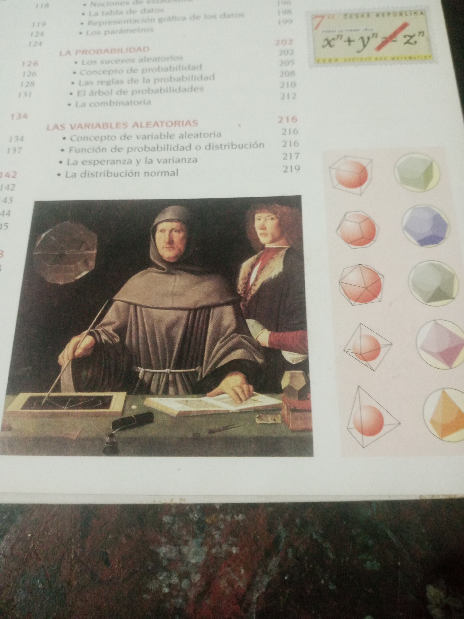 The image displays a page from a book or publication. On the left side, a painting depicts two figures in historical attire. The foreground figure, a man with a tonsure, wears a monk's habit and is engaged in drawing or writing on a surface with a stylus. Behind him, another figure, possibly a young man, stands with a serious expression. To the right of the painting are lists of chapter titles and page numbers, indicating a text related to mathematics or statistics, with terms like "Probabilidad" (Probability) and "Variables Aleatorias" (Random Variables). Further to the right, a series of geometric shapes, rendered as spheres within wireframe structures and polyhedra, are displayed. At the top right corner, a postage stamp with the text "CESKÁ REPUBLIKA" and a mathematical equation is visible. There is no information in the image that relates to the city of Santa Ana, Venezuela.