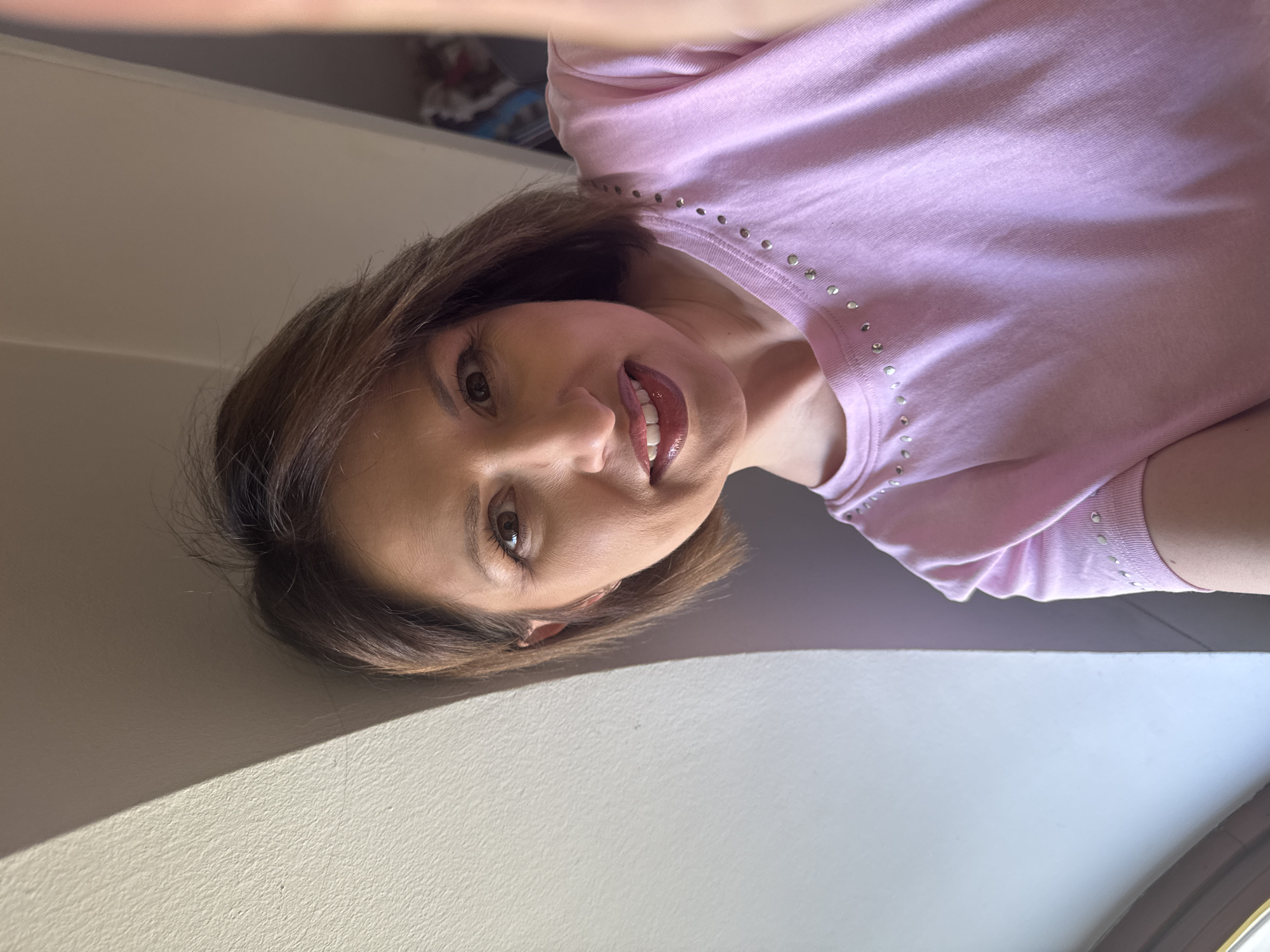 A person with brown hair is posing for a selfie. She is wearing a light pink shirt with studs around the neckline. The location appears to be indoors, possibly in Sofia, Bulgaria. A wall is visible in the foreground, and there is some light streaming in from a window.