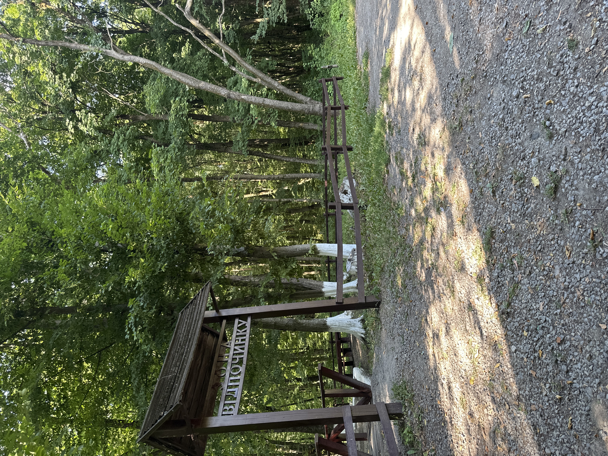 The image depicts an outdoor scene in a wooded area. In the foreground, a gravel path is visible, with patches of grass and fallen leaves. Sunlight casts shadows across the path.
To the left, there is a wooden archway with a wooden roof. Suspended from the arch is a wooden sign with text in Cyrillic. The sign appears to read "ВОДА ВІДПОЧИНКУ" which translates to "Water of Relaxation". The archway is supported by two wooden posts. To the left of the arch, a wooden bridge or walkway structure is partially visible.
Further into the scene, behind the archway, is a wooden fence. Several trees with white-painted bases are visible behind the fence. The area behind the fence is densely wooded with lush green foliage. The overall impression is of a natural setting, possibly a park or recreational area, with a marked entrance. The image appears to be taken during daylight.