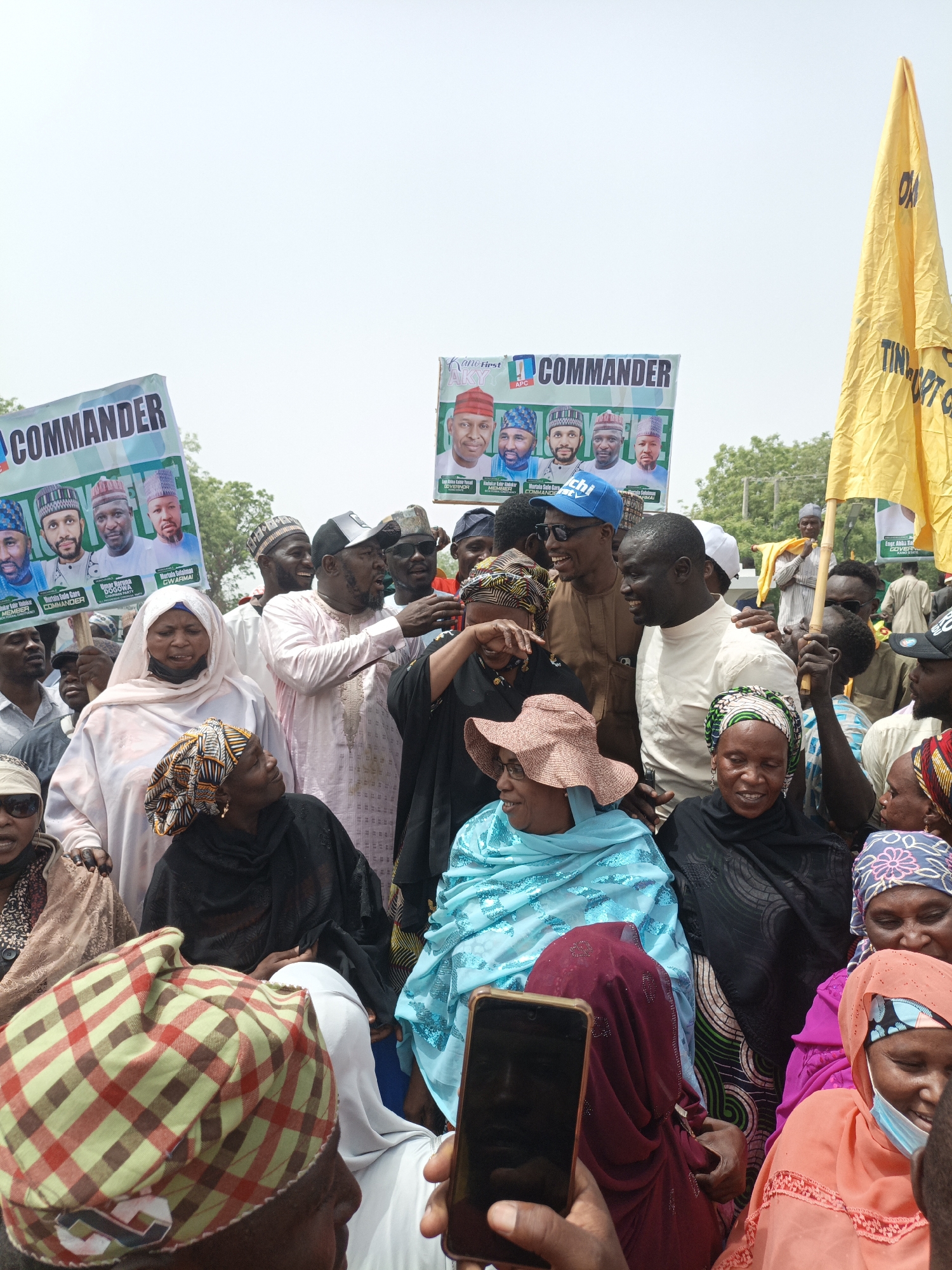 A vibrant outdoor gathering, likely a political rally, is captured in this daylight image. A large crowd of people, primarily adults, fills the frame, with both men and women dressed in a mix of traditional and casual attire. Many women wear colorful headscarves or hijabs, while men are seen in caps and robes or t-shirts.
Several individuals appear to be central figures, engaged in conversation and interaction with the crowd. A man in a brown shirt and blue "Chi" cap is seen smiling and leaning into the group, interacting with a woman in black. Another man in a white t-shirt is smiling broadly, surrounded by people. The overall atmosphere seems lively and engaged, with many smiling faces visible, indicating enthusiasm.
Prominent in the scene are several political campaign banners. Two large banners feature the word "COMMANDER" and display portraits of multiple men, likely candidates. Visible text on these banners includes "MEMBER COMMANDER," "Murtala Sule Gare," "DOGOWA," "Murtala Sulaiman," and "GWARMAI." Another banner on the right side features "Engr. Abba Kabir" and the word "GOVERNOR," along with "APC," indicating the All Progressives Congress political party. A large yellow flag, partially visible on the far right, also carries text, possibly "TINUBU GANI" or similar political slogans. A smartphone is visible in the foreground, held by someone capturing the event.
The setting appears to be an open public space under a clear, bright sky, suggesting a sunny day. Trees are visible in the background, further confirming an outdoor location.