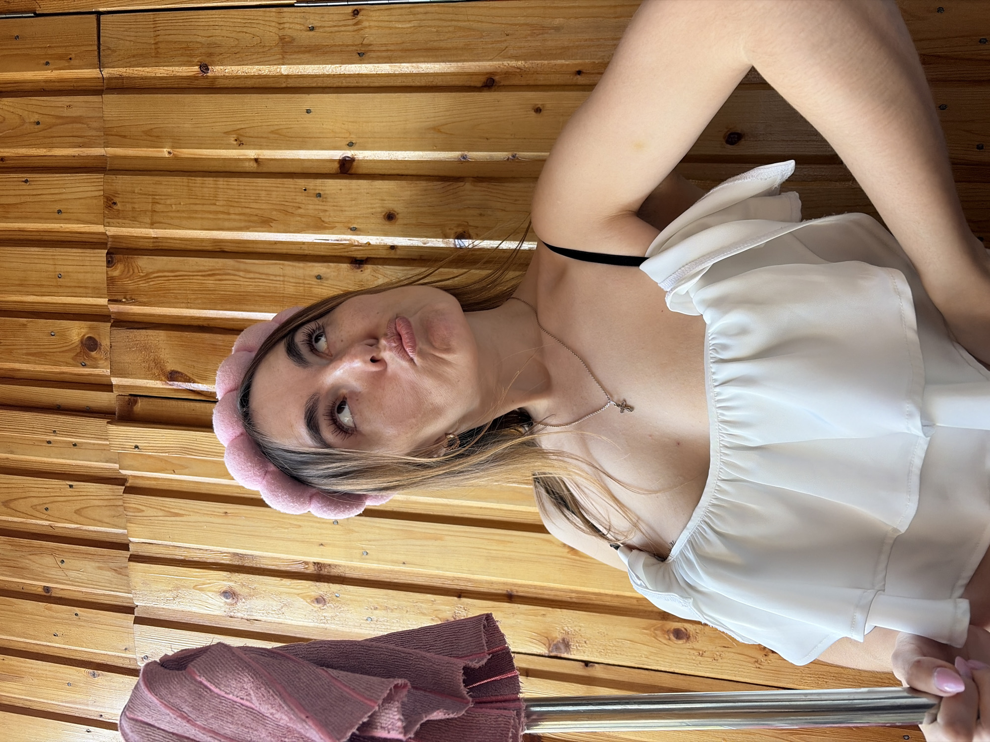 The image shows a young woman with long brown hair, wearing a pink flower-shaped headband and a white ruffled top. She has light skin, brown eyes, and is making a pouting face. She is wearing a silver necklace with a cross pendant. The background is a wooden wall made of horizontal planks. A pink striped towel or blanket is visible in the lower left corner of the image, and a silver metal bar is in the lower right corner. There is no indication of Izhevsk, Russia, or any other city.