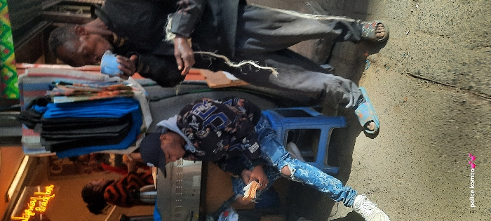 The image depicts a daytime street scene on a commercial thoroughfare in Nairobi, Kenya. In the foreground, two males are present. On the left, a younger male sits on a blue plastic stool, wearing a black baseball cap, a dark patterned hoodie, ripped blue jeans, and white sneakers. He holds a thick stack of paper currency in both hands. To his right, an older male stands, dressed in a dark long-sleeved top and dark sweatpants with a light stripe. He drinks from a light blue cup with his right hand, while a long, light-colored fibrous material hangs from his left hand. He wears a dark sandal on his right foot and a light blue slider on his left.
The background features a commercial establishment with an illuminated sign displaying "California - snack bar." A female figure, wearing a striped top and with her hair tied back, is partially visible behind a metallic counter inside this establishment. Adjacent to the establishment, a market stall setup is discernible, featuring various textiles and fabrics hanging vertically. The ground is a coarse, uneven concrete or asphalt surface. The scene is brightly lit by natural sunlight.
