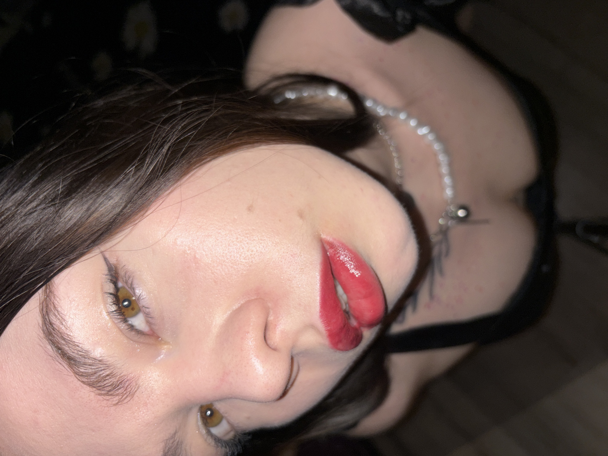 A close-up, tilted portrait of a young woman with fair skin and dark brown hair. Her eyes are hazel, and she wears dark eyeliner. Her lips are painted with glossy red lipstick. She has a silver chain necklace. A black garment, possibly lingerie, is visible on her body. There is a dark, indistinct background. There is no indication of the location being in Perm, Russia, based on the image.