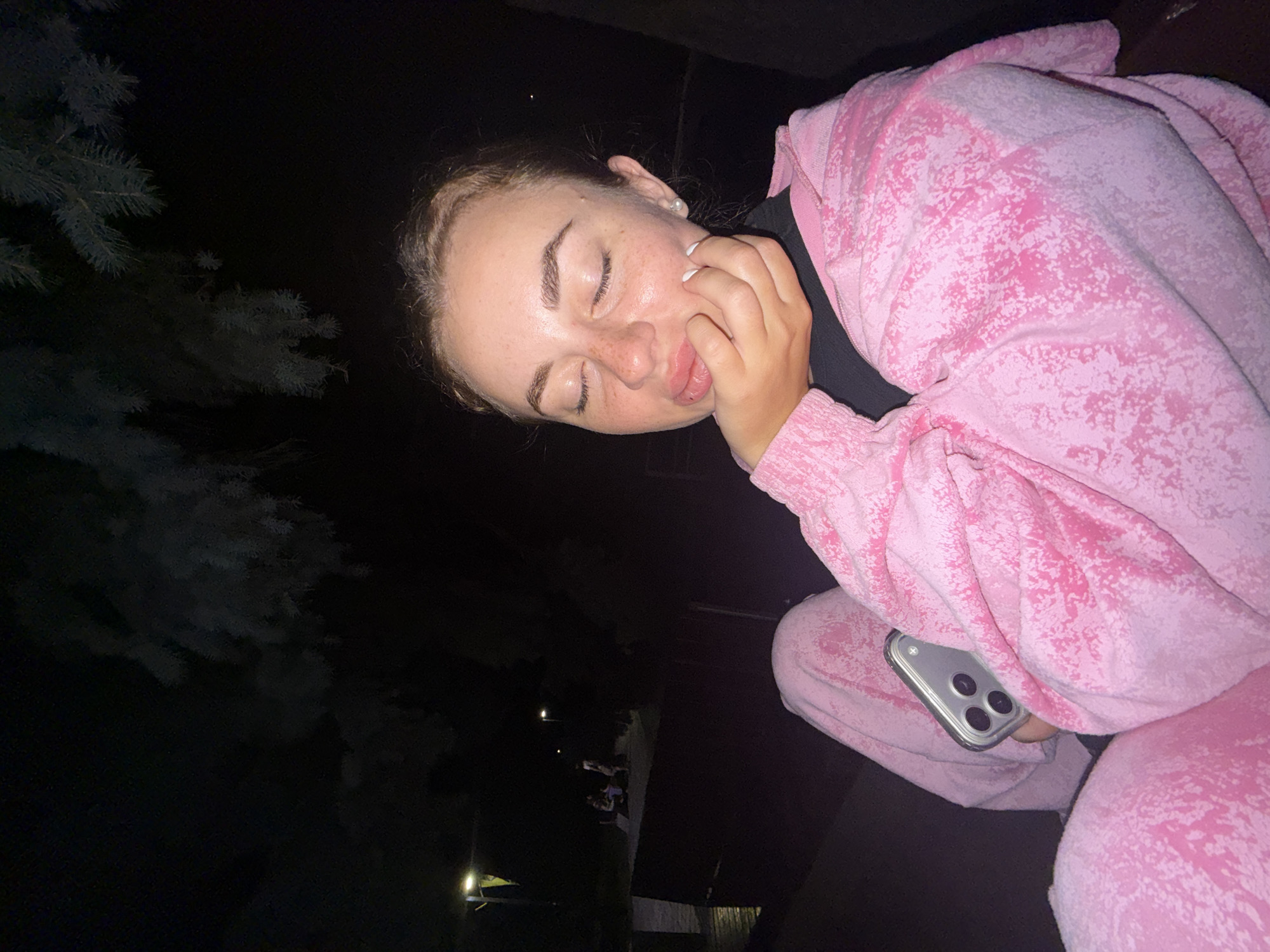 A young woman, with her eyes closed and a playful expression, is pictured at night in Dnipro, Ukraine. She is wearing a pink tie-dye loungewear set, with a matching hooded jacket and pants. Her hand is touching her face, and she has a pearl earring in her left ear. A cell phone is peeking out from under her clothing, with its camera lenses visible. The background is dark, with the silhouette of a tree on the left side and a distant light in the lower right corner. There are also a few other figures faintly visible in the distance, suggesting the presence of other people in the scene. The overall mood of the image is casual and relaxed, with the woman appearing to be in a moment of quiet enjoyment or jest.