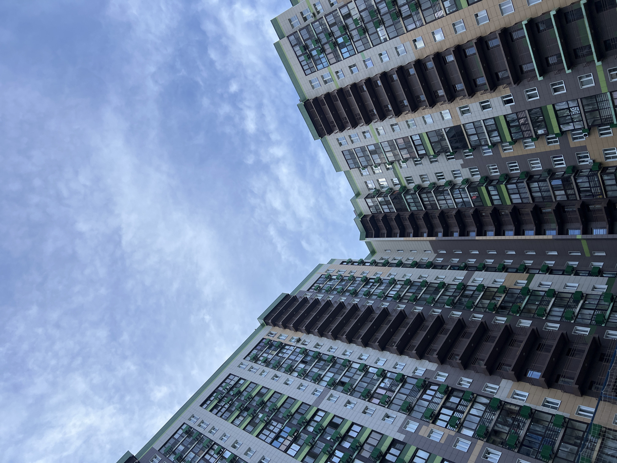 A low-angle shot captures two apartment buildings extending upwards to a sky filled with thin clouds. The buildings are multi-storied with numerous windows and balconies. The architecture features a mix of neutral tones, including white, gray, and brown, with accents of green. The windows reflect the sky, and the balconies appear dark. The angle of the shot suggests the photographer is standing directly below the buildings, creating a converging perspective. This image is likely taken in Khimki, Russia, given the distinctive architectural style and urban environment.