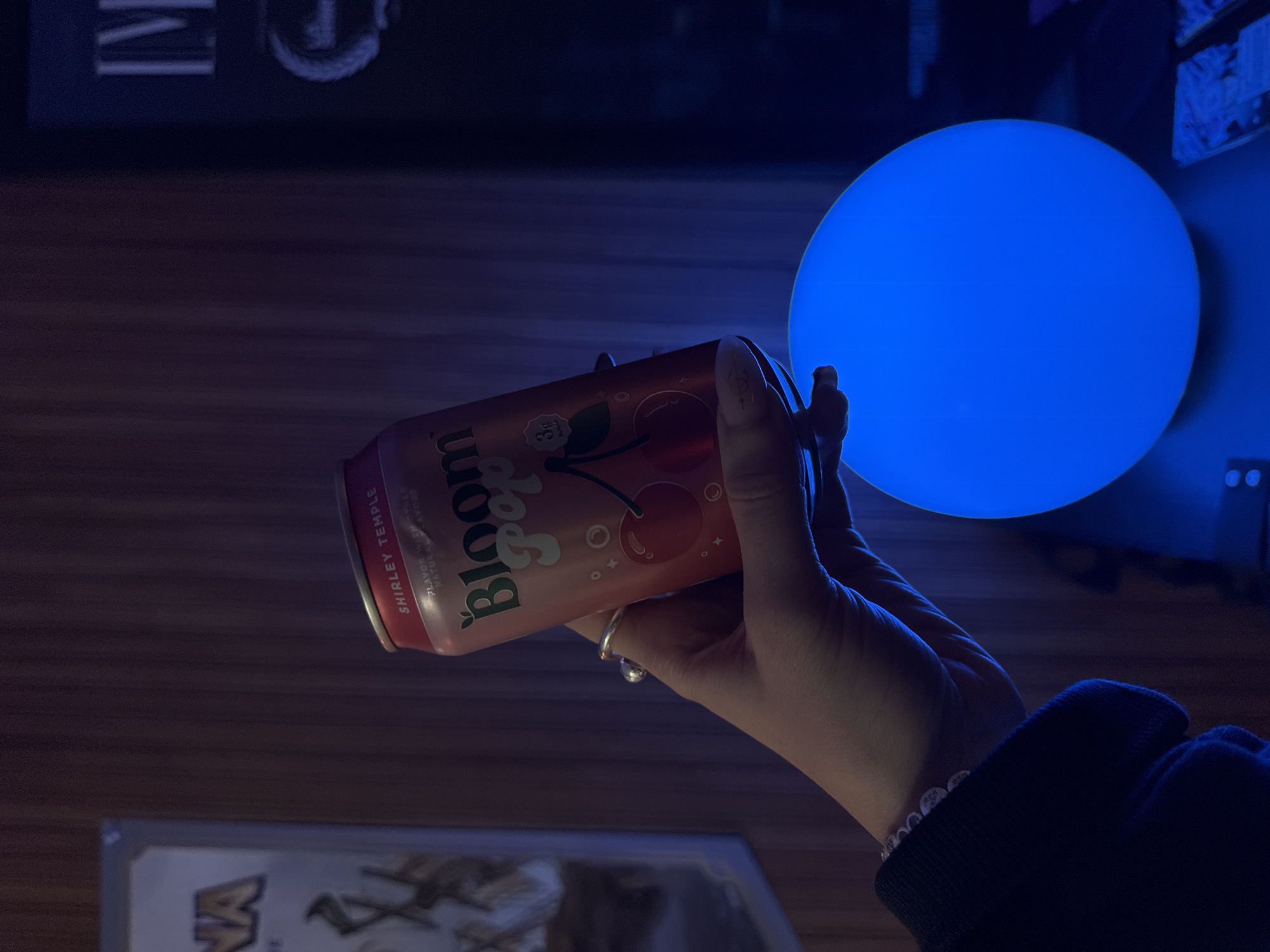 The image depicts a hand holding a can of "Bloom Pop" Shirley Temple soda in front of a blue orb-shaped light source. The background includes a dark wooden surface and glimpses of what appears to be a television or monitor. The lighting is predominantly blue and dim, creating a somewhat moody atmosphere. The details are insufficient to determine the exact location in Los Angeles.