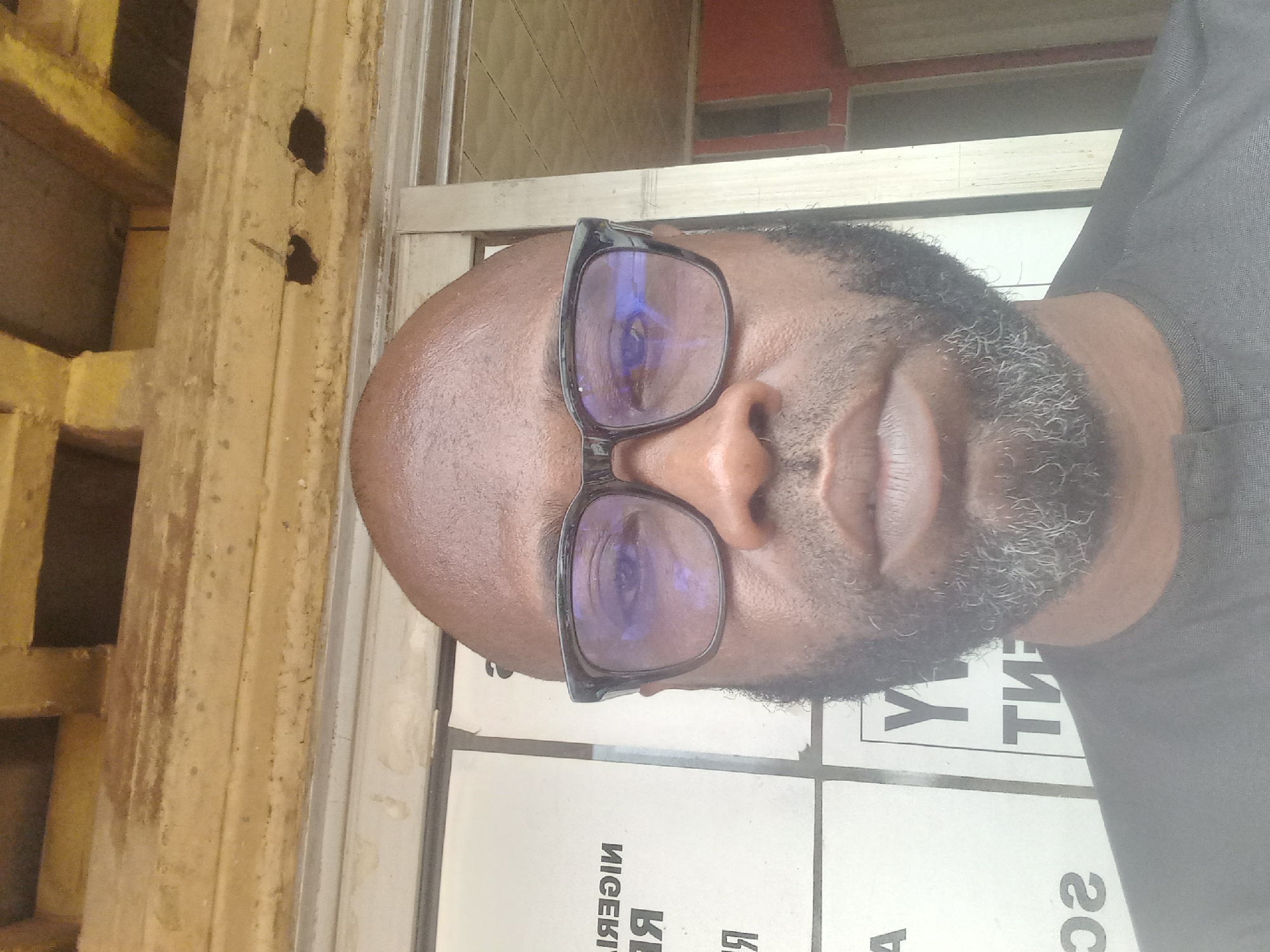 A close-up, slightly angled portrait of a man with a bald head, dark skin, and a greying beard. He is wearing glasses with a black frame. The reflection in his glasses reveals a blurred interior space. He appears to be positioned in front of a window, as evidenced by the light and the visible window frame. Behind the window, through which light is penetrating, are white rectangular panels with black text. The text is partially obscured and in reverse. To the left of the window is a wooden structure with holes. The man's shirt, visible to the right, is dark in color. The location is suggested to be in Sardauna, Nigeria, but there are no identifiable landmarks or features within the frame to confirm this.