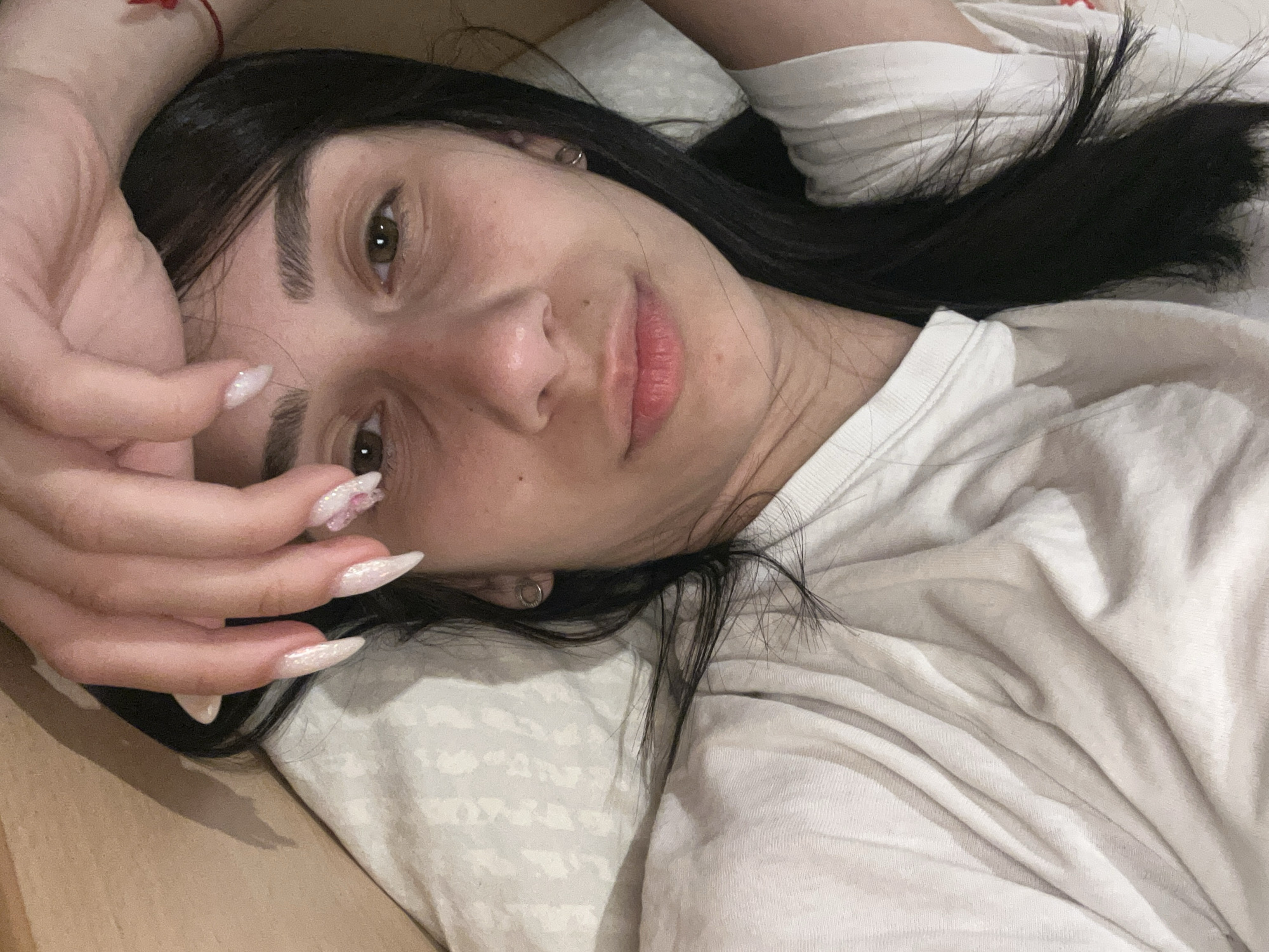 A young woman is lying down, possibly on a bed, wearing a white t-shirt. She has long, dark hair and is looking at the camera. Her hand is raised near her face, displaying long, pointed nails with a decorative pattern. The background includes a pillow and what appears to be a wooden surface. The location of the photo in Plovdiv, Bulgaria is not discernible from this image.