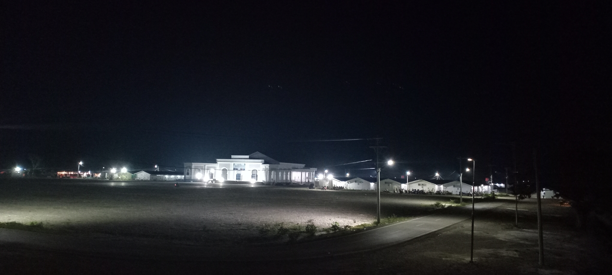 The image depicts a nighttime scene in Meureudu, Indonesia, featuring a large, open field under a dark sky. In the mid-ground, a prominent, brightly illuminated, multi-story building with classical architectural elements, including columns and a central balcony, stands out with its light-colored facade. To the right of this central structure, a row of approximately eight smaller, white, single-story buildings with pitched roofs are also illuminated. Several individuals are observed in small groups near these smaller buildings. To the far left, additional smaller, illuminated structures are visible.
A paved road or pathway curves from the lower right foreground towards the center, bordered by at least three operational streetlights mounted on utility poles. Overhead power lines span the scene. The foreground consists of a vast, flat, unpaved or grassy area, partially lit by the distant lights. The overall environment suggests an institutional or public complex, possibly with temporary facilities, at night.
