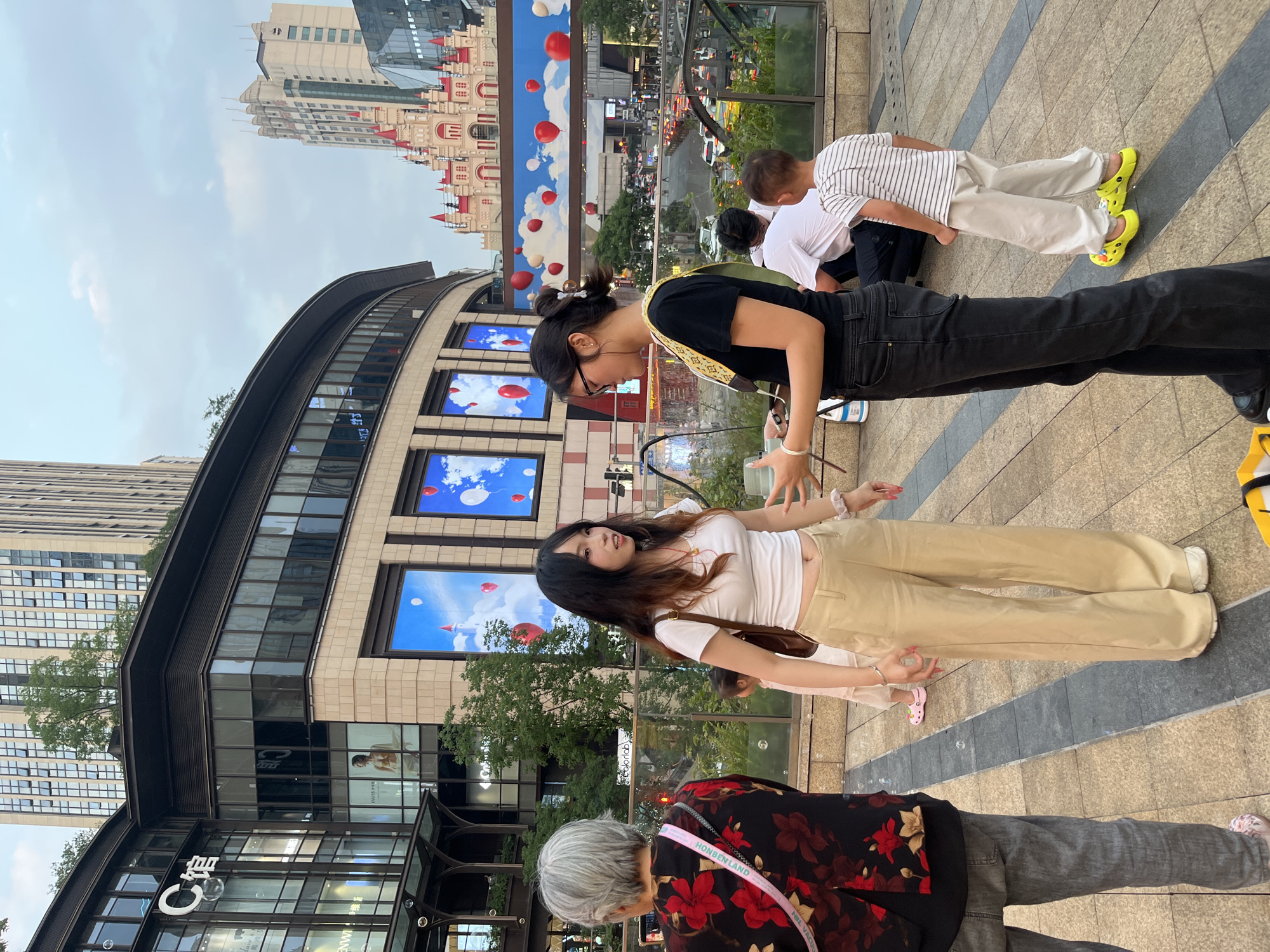 This image depicts an outdoor pedestrian area at Raffles City Chongqing's C Hall in Yuzhong District, China, with Changjiahui Shopping Park visible across the river in the background. The scene unfolds under an overcast sky during late afternoon or early evening.
In the foreground, an elderly woman with short gray hair, dressed in a black shirt with red floral patterns and gray pants, faces away from the viewer. She carries a pink and white shoulder bag with "HONBENLAND" visible on the strap. To her right, a young woman with long, dark hair and lighter ends, wearing a white crop top, beige wide-leg pants, and white shoes, stands facing right, engaged in conversation. She wears a red pendant necklace and carries a brown shoulder bag. Opposite her, a second young woman with dark hair tied back, wearing black glasses, a black t-shirt with a patterned shoulder strap, black jeans, and black shoes, holds a white object in her left hand and gestures with her right. Bubbles float in the air around the individuals.
In the mid-ground, a man in a white t-shirt and dark pants is seated on the ground with his back to the viewer. To his right, a young boy, also seen from behind, wears a striped t-shirt, light pants, and bright neon yellow Crocs-style shoes. A small child in a light dress and pink shoes is partially visible behind the first young woman.
The background features the curved glass and stone facade of Raffles City Chongqing's C Hall ("C馆") on the left, displaying large windows and a prominent "C馆" logo. Large digital display screens on a building in the mid-ground showcase images of a blue sky with white clouds and red balloons, alongside a smaller depiction of a castle. Further in the background, a distinctive red-roofed, multi-turreted castle-like structure, part of Changjiahui Shopping Park in Nan'an District, is visible. Multiple modern high-rise buildings and skyscrapers dominate the distant skyline. Below the pedestrian level, a busy multi-lane road with vehicles is visible through glass railings. The pedestrian area is paved with gray and dark gray striped tiles.