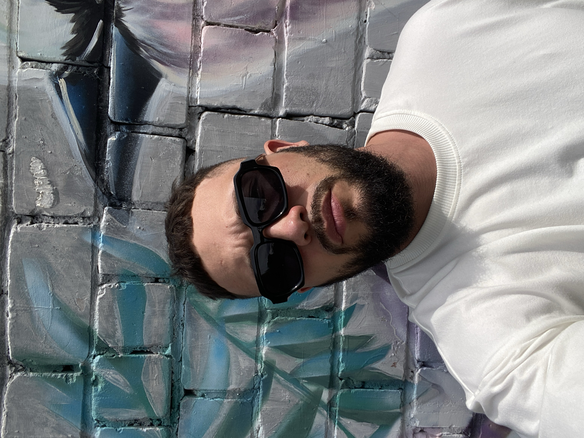 A close-up, chest-up portrait of a man wearing sunglasses and a white sweatshirt, standing against a graffiti-covered brick wall. The man has dark hair, a beard, and is looking directly at the camera. The graffiti on the wall features abstract shapes and colors, primarily in shades of blue, teal, gray, and pink. The lighting suggests it is daytime, with harsh shadows. The specific location of Chaikovsky, Russia, is not determinable from the image itself.