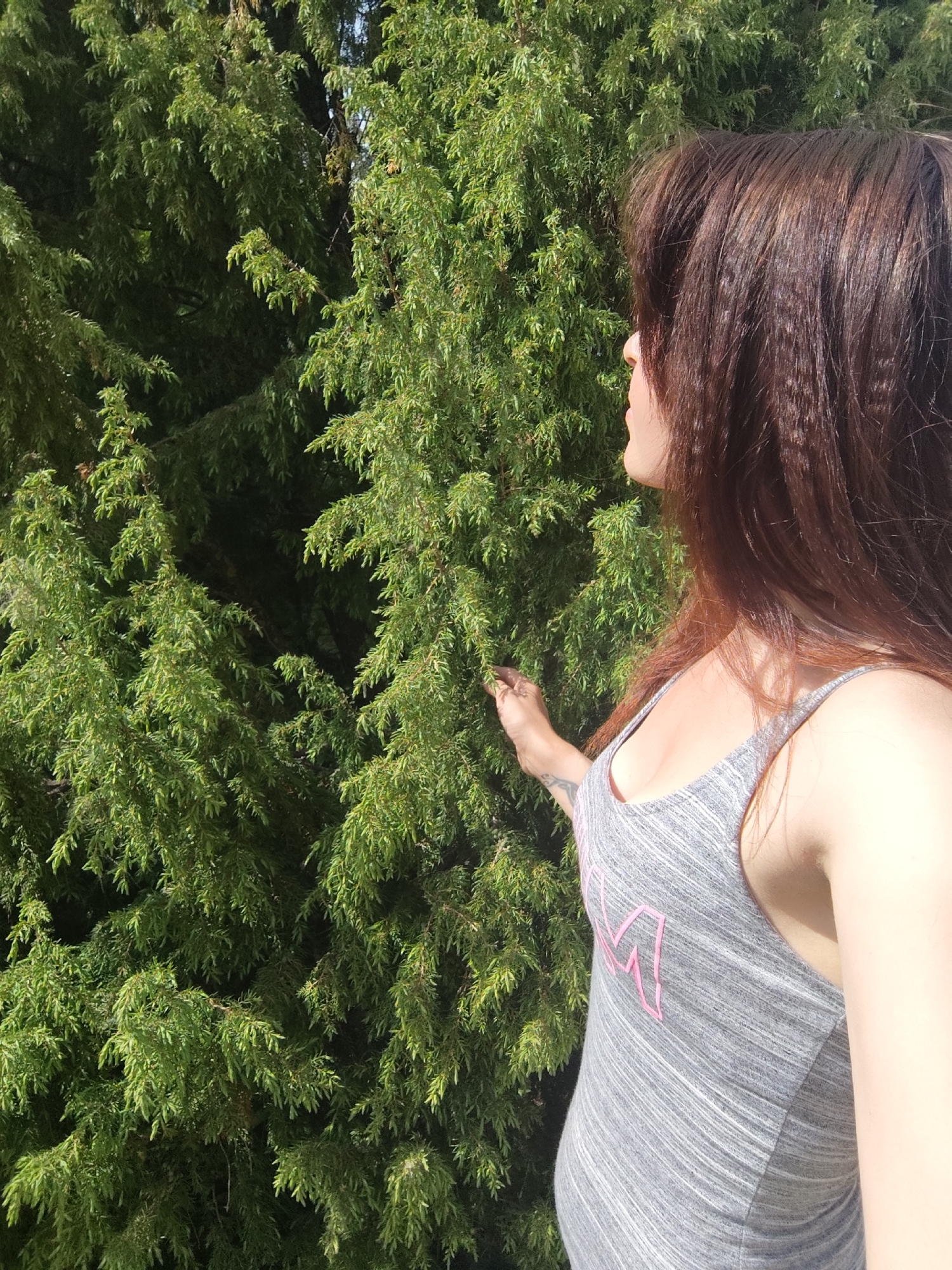 A woman in a grey tank top stands next to a green coniferous tree. The woman has long brown hair and is looking to the left of the frame. Her right hand is extended, and her fingers appear to be touching the tree. The background consists of dense green foliage. The photo does not provide sufficient information to identify the precise location within the city of Фоки, Russia.