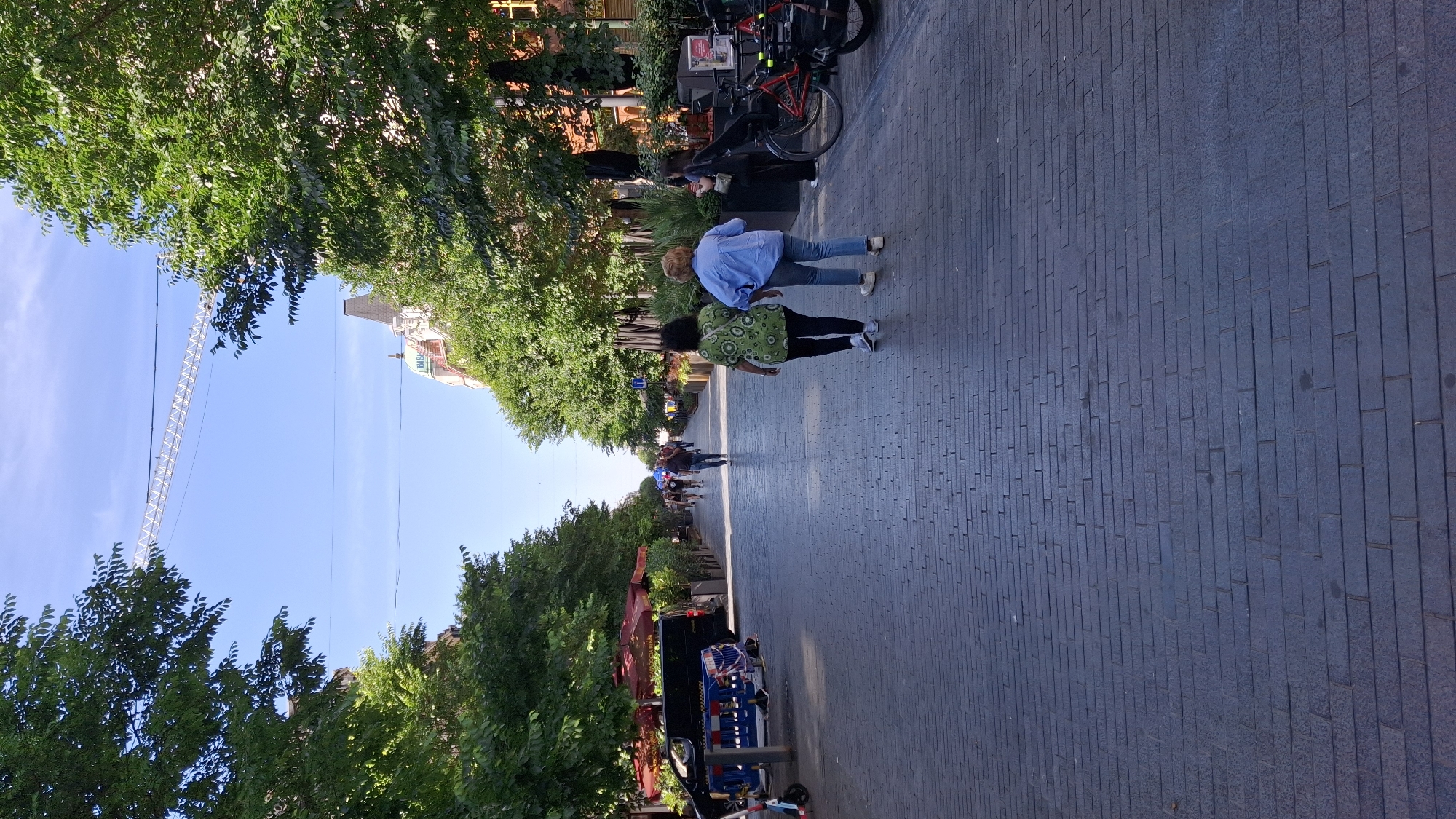 A pedestrian street in an urban setting is depicted under a clear blue sky. The street is paved with dark gray bricks and lined on both sides with trees that create a canopy. In the foreground, two women walk away from the camera. The woman on the left wears a green, patterned short-sleeved shirt and black pants, while the woman on the right wears a light blue long-sleeved shirt and blue jeans. In the middle ground, other pedestrians are walking along the street. On the left side of the street, there are red umbrellas, a black van, and some barriers. On the right side, bicycles are parked, and there are outdoor seating areas for businesses. In the background, buildings with architectural details are visible, and a large construction crane extends across the sky. The overall scene suggests a lively commercial or recreational area.