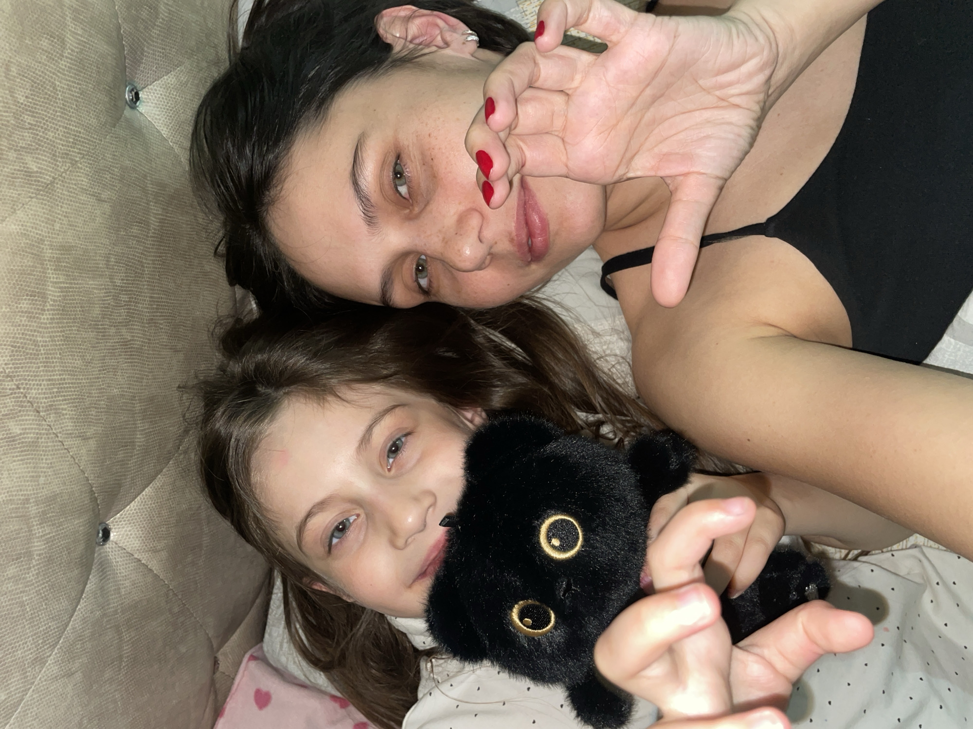 A woman and a young girl are lying in bed together. The woman is wearing a black tank top and has red nail polish. She is making a hand gesture towards the camera. The girl is holding a black plush panther toy. They are in a bed with a headboard. The location cannot be determined from the image.