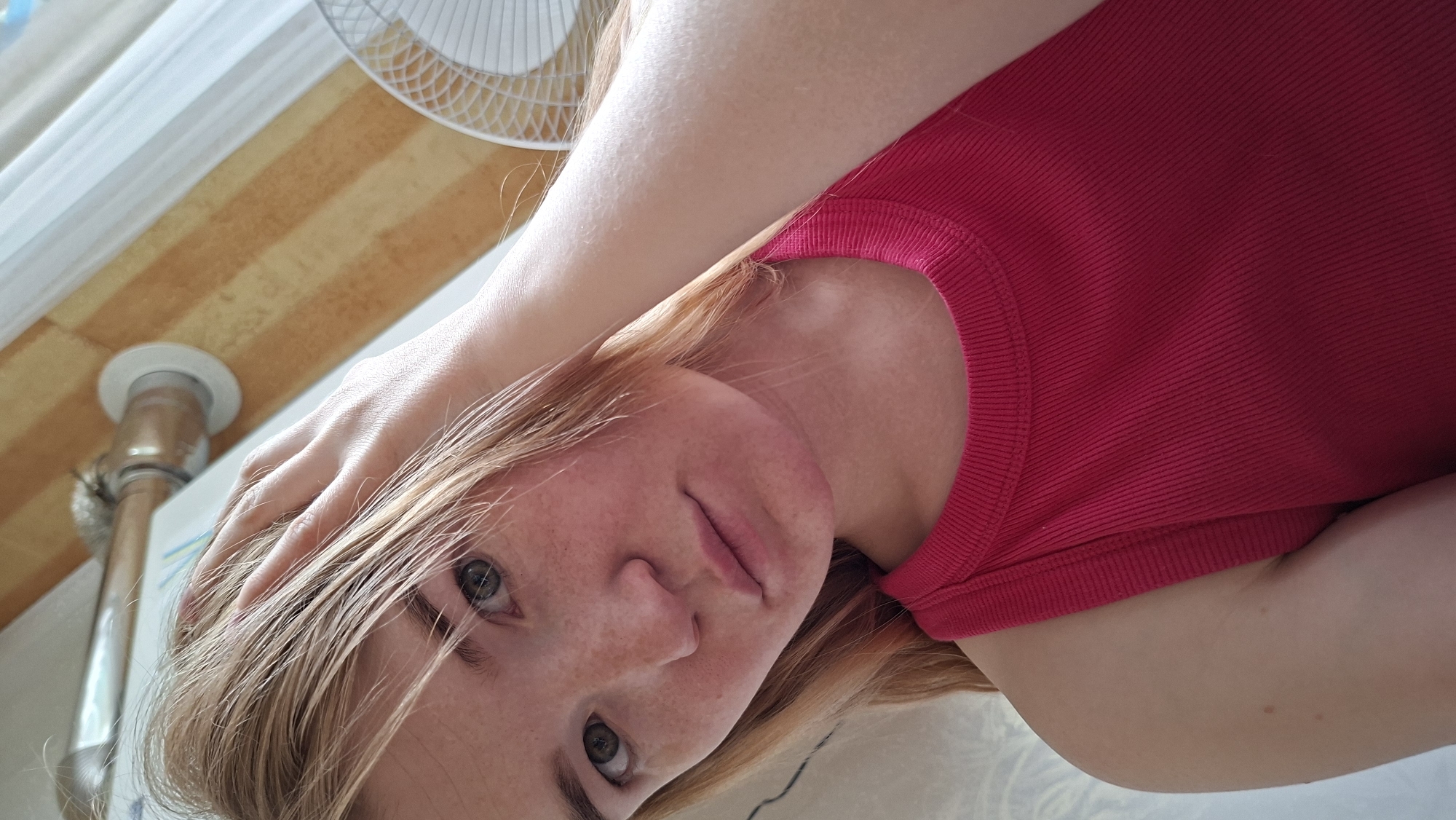 A young person, likely female, with fair skin, light brown or blonde hair, and blue-green eyes, is depicted lying down, looking directly up at the camera. Her face shows scattered freckles, and she wears a bright, ribbed red sleeveless top. One hand is casually placed on the side of her head, with fingers resting in her hair.
The setting appears to be indoors, likely a bedroom or a similar personal space, viewed from an overhead perspective. The background features a distinctive striped pattern on what seems to be a wall or ceiling, composed of alternating light brown and beige/yellow bands. A portion of a white ceiling fan is visible in the upper right corner, and a light-colored curtain or blind can be seen along the left edge. Towards the bottom left, a metallic pipe, possibly part of a radiator or furniture, is visible, secured by a white circular fixture at its base.
The lighting is soft and natural, suggesting it is daytime. The person's expression is calm and neutral, conveying a sense of relaxation. There is no visible text within the image.