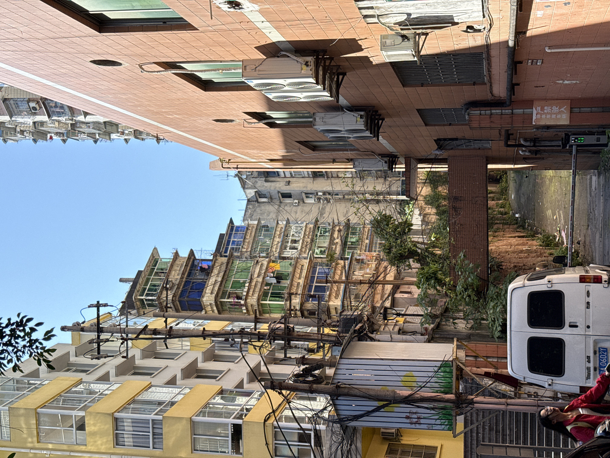 The scene depicts a bustling urban environment in Beibei, Chongqing, China. It is a sunny day. A dense network of overhead power lines and cables crisscrosses between buildings. The buildings themselves display diverse architectural styles, ranging from older structures with visible weathering and eclectic window designs to more modern apartment blocks. A white delivery van is parked on a narrow street. A woman in a red jacket stands near the van and is looking at her phone. Signs in Chinese script are visible on the buildings.