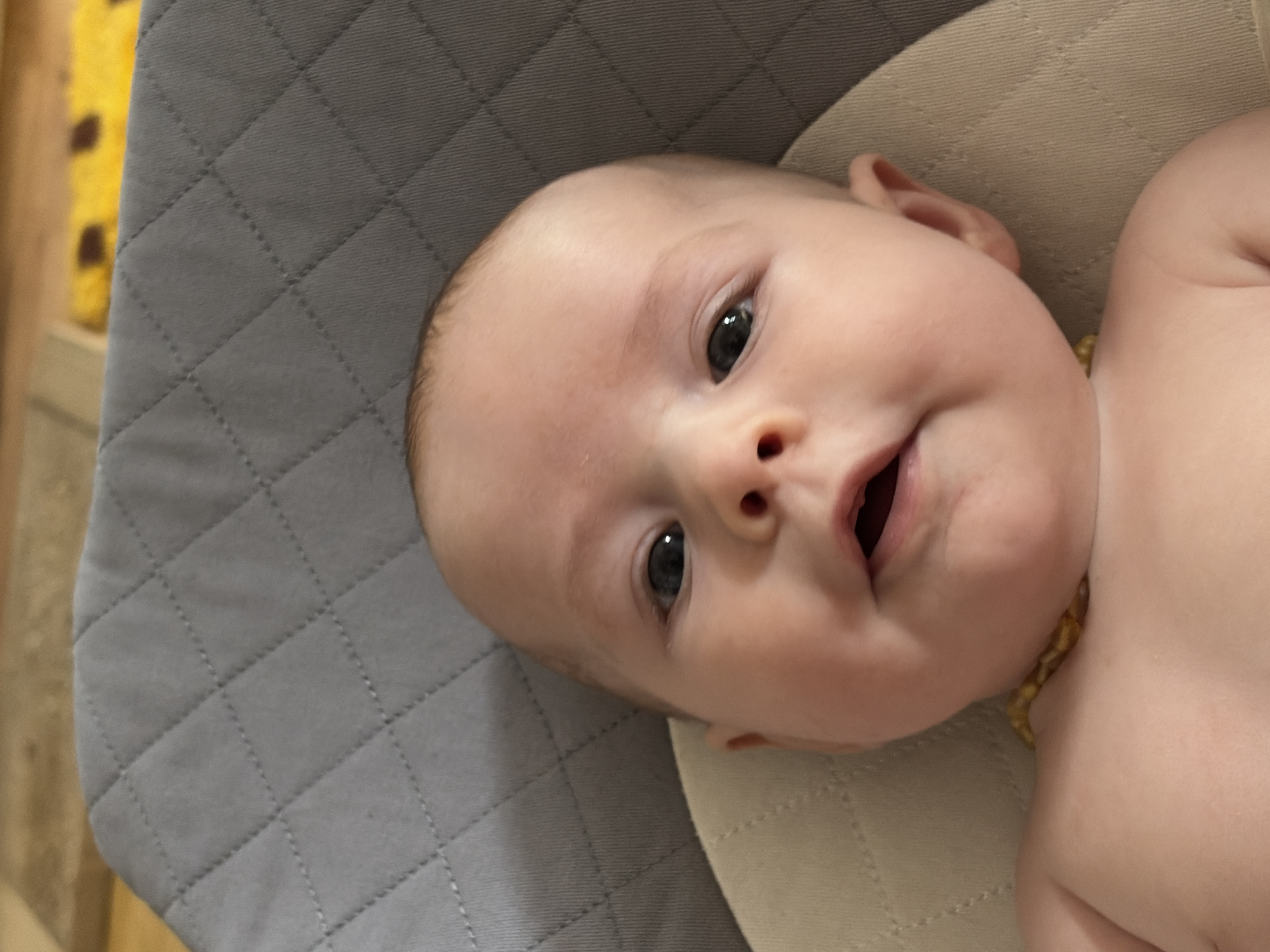 A close-up shot depicts a baby lying on a gray quilted surface with a beige section visible near the baby's head. The baby's face is the focal point, with the head turned slightly towards the camera. The baby has fair skin, dark eyes, and an open mouth. A necklace made of small amber beads is visible around the baby's neck. The setting appears to be indoors. It is not possible to determine if this photo was taken in Chaikovsky, Russia.